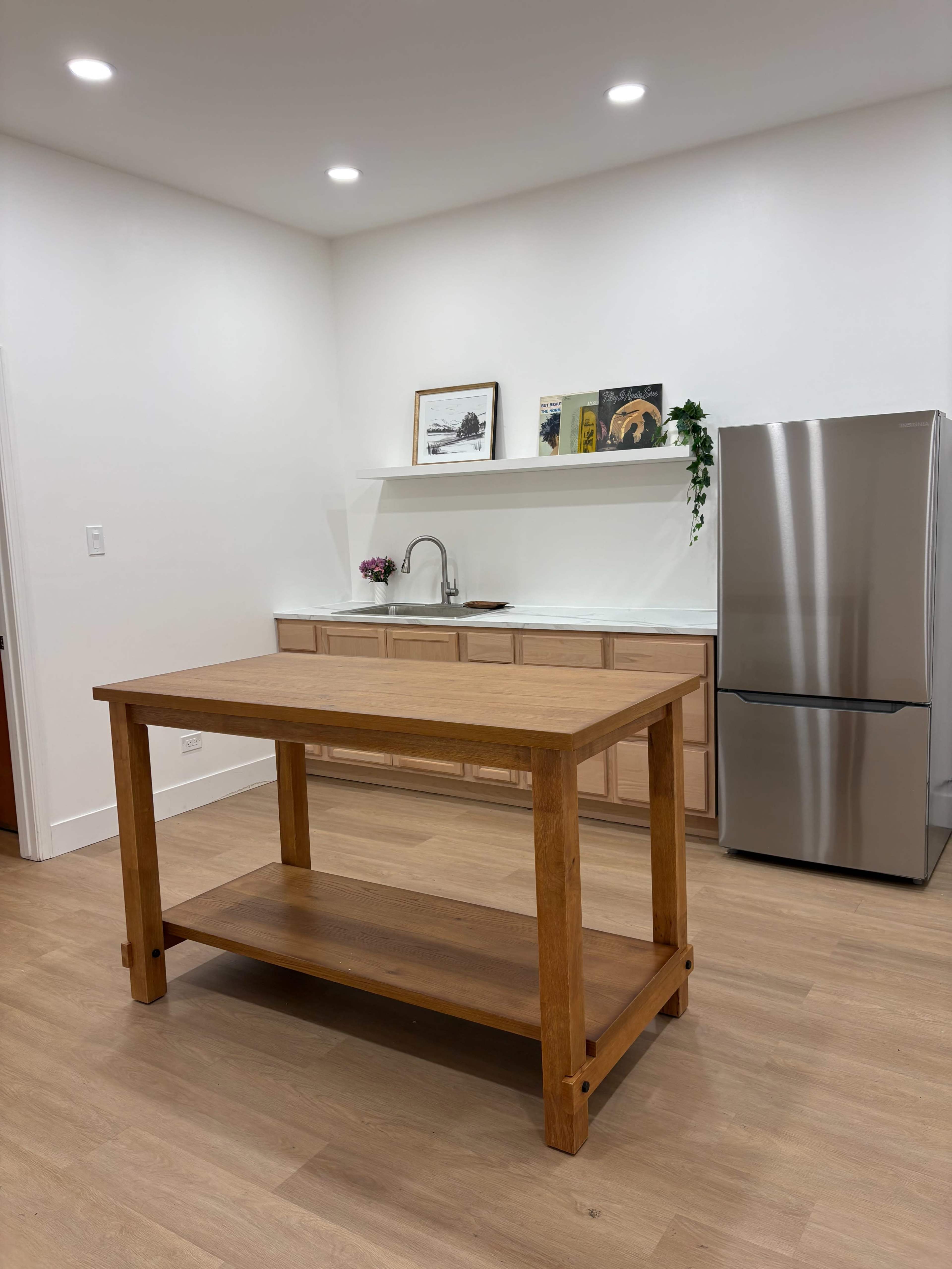 The image shows a modern kitchen with a wooden island in the center, stainless steel refrigerator on the right, and light-colored cabinetry in the background.