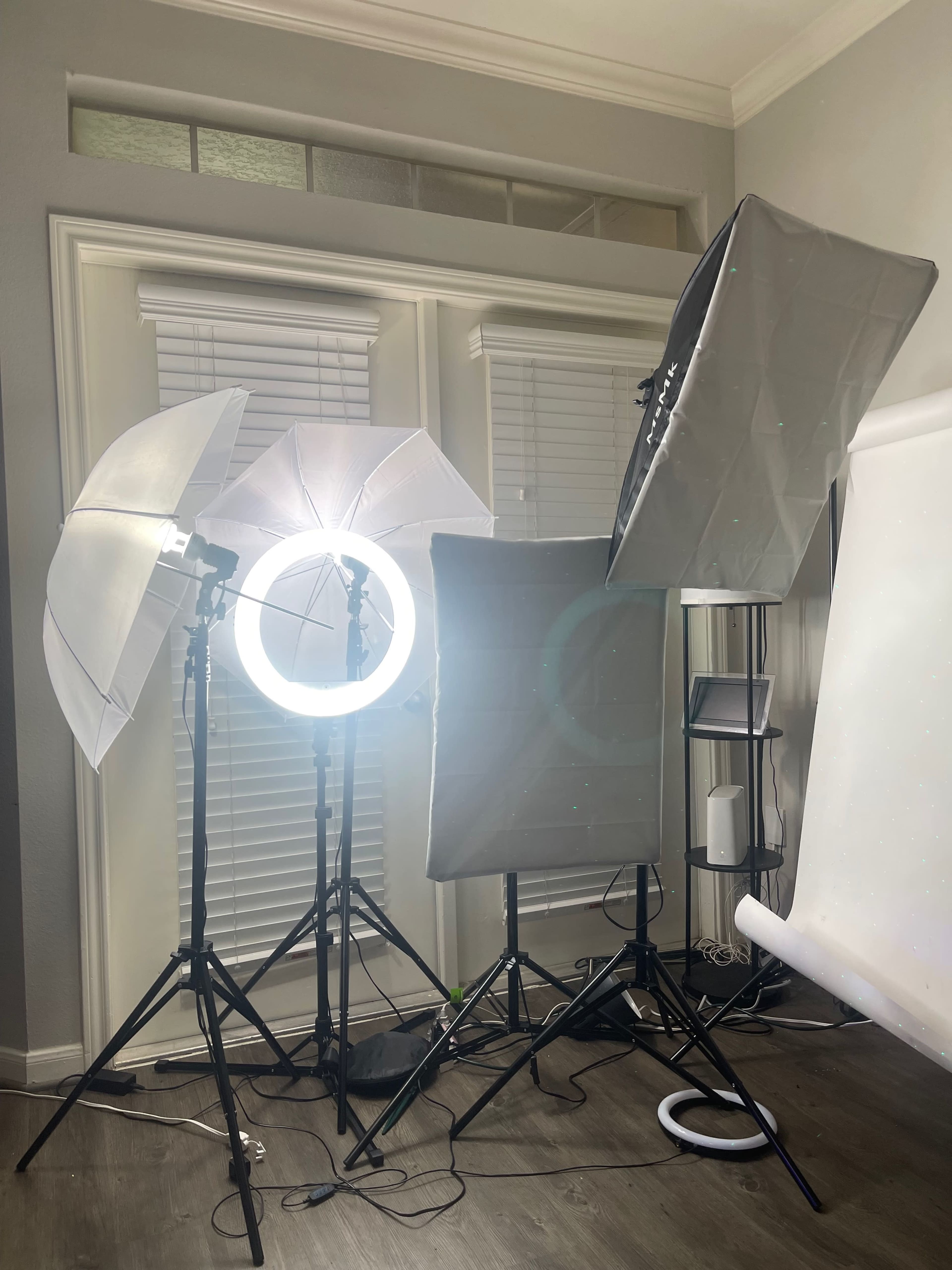 A photography setup featuring multiple light sources, including softboxes and a ring light, arranged around a backdrop in a room with white blinds.