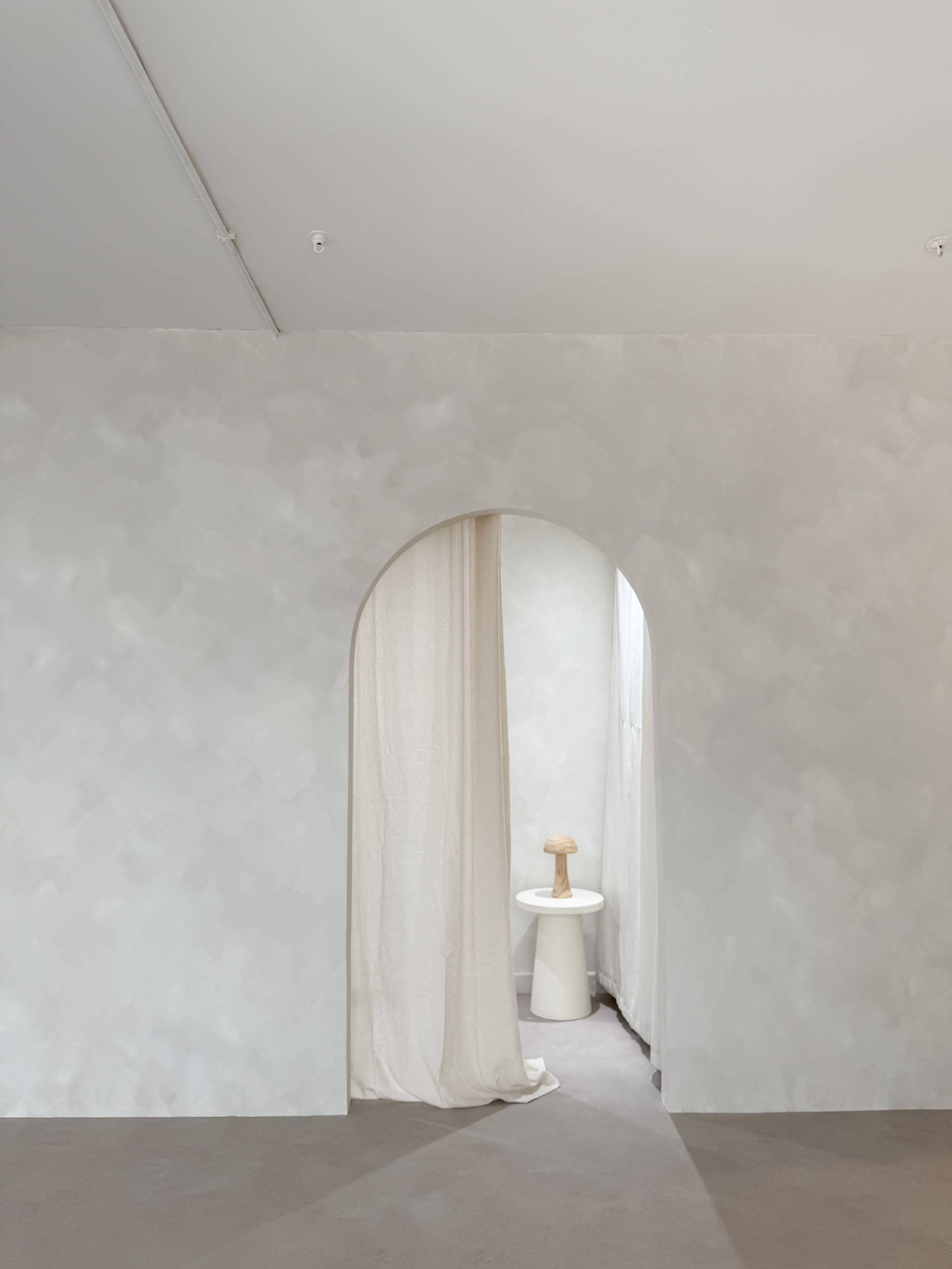 A beige wall with a rounded archway leads to a softly lit area featuring a small stool and a white curtain.