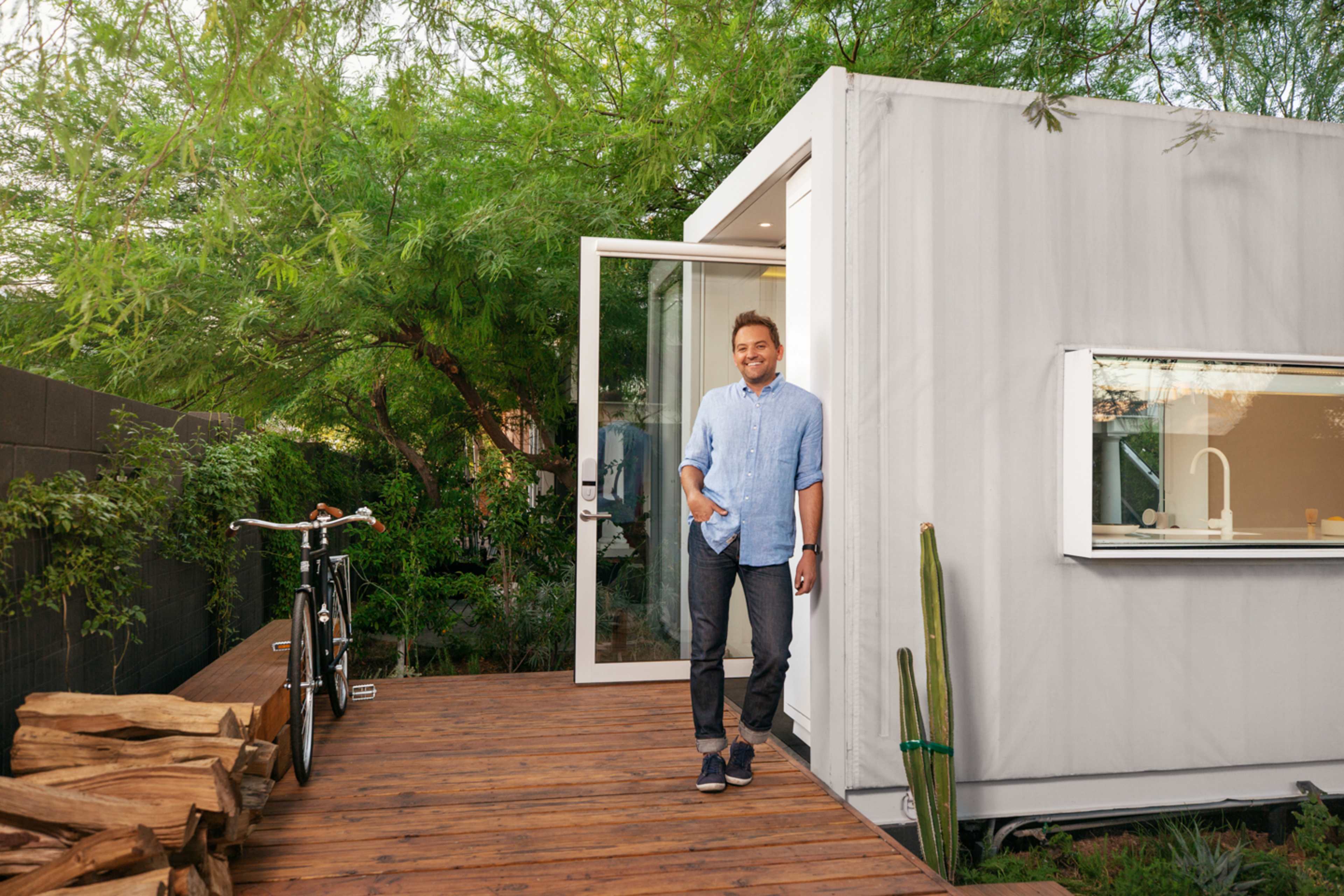 A man stands on a wooden deck next to a modern, minimalist house surrounded by greenery and a bicycle leaning against the wall.