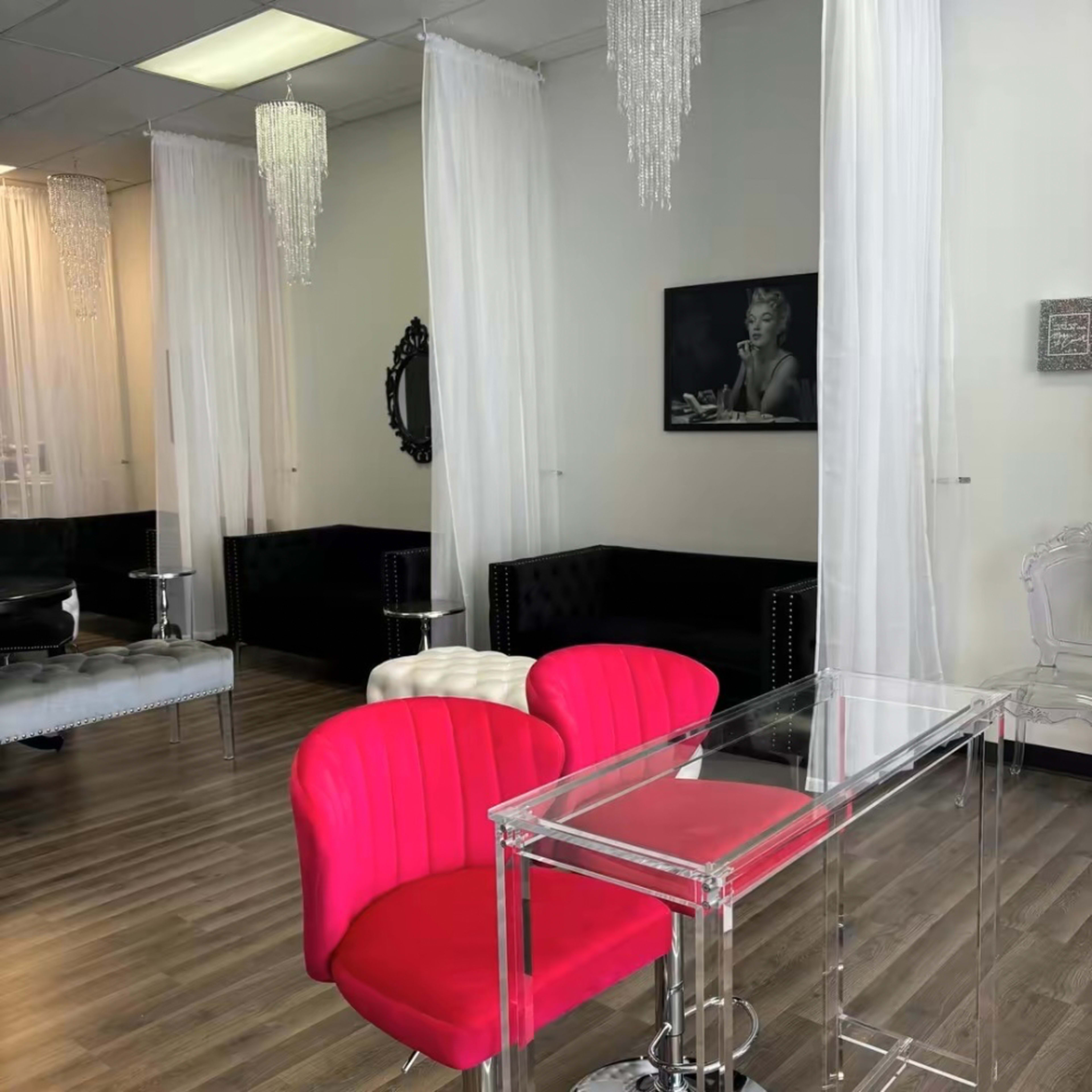 The interior of a salon features pink and clear chairs, black couches, and sheer white curtains, with chandeliers hanging from the ceiling.