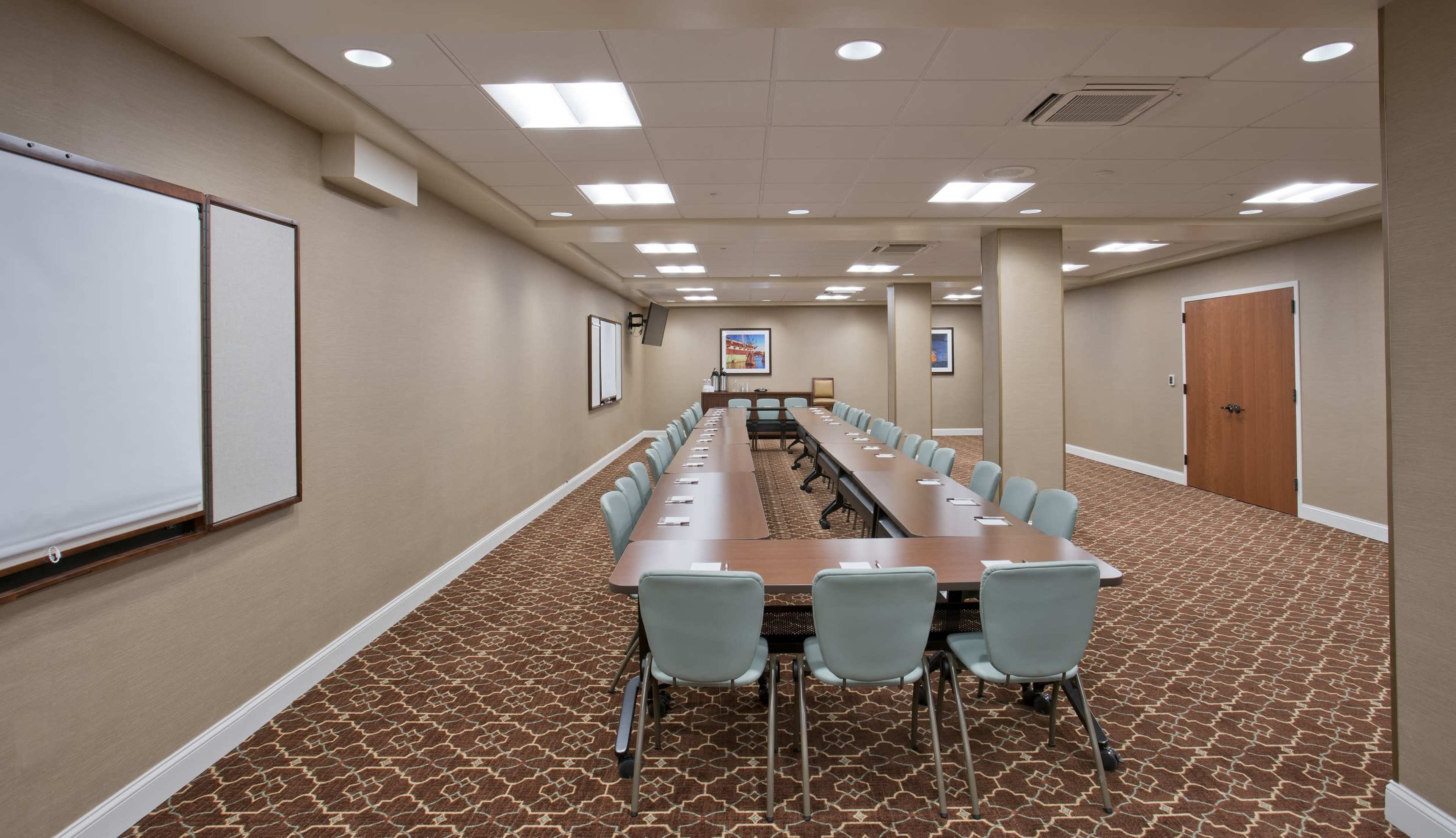 A conference room features a long table with mint green chairs arranged in two parallel rows, flanked by whiteboards and equipped with wall-mounted screens.