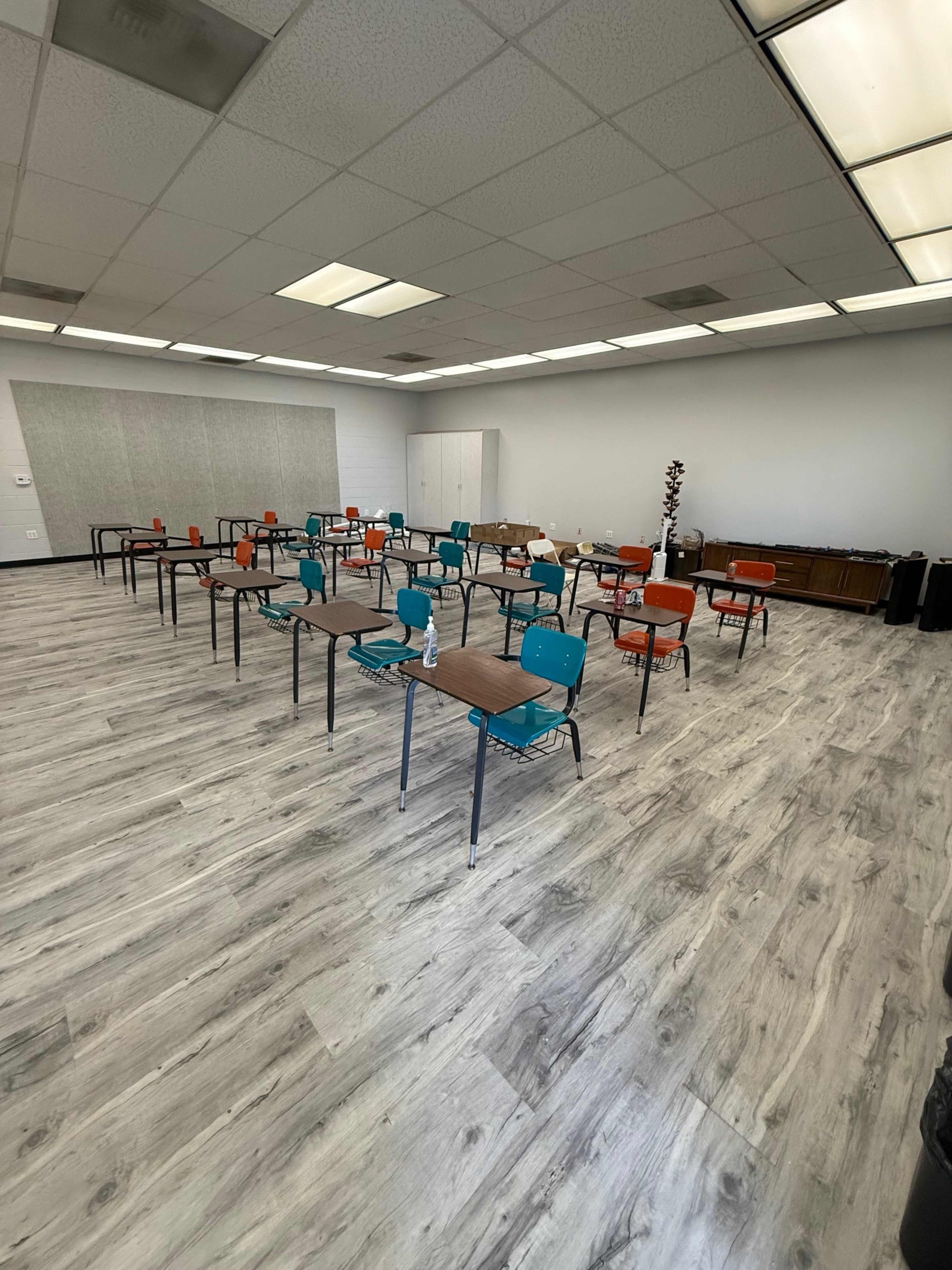 The image shows a classroom with several tables and chairs arranged in a grid pattern, featuring turquoise and orange seats, and a light-colored wooden floor.