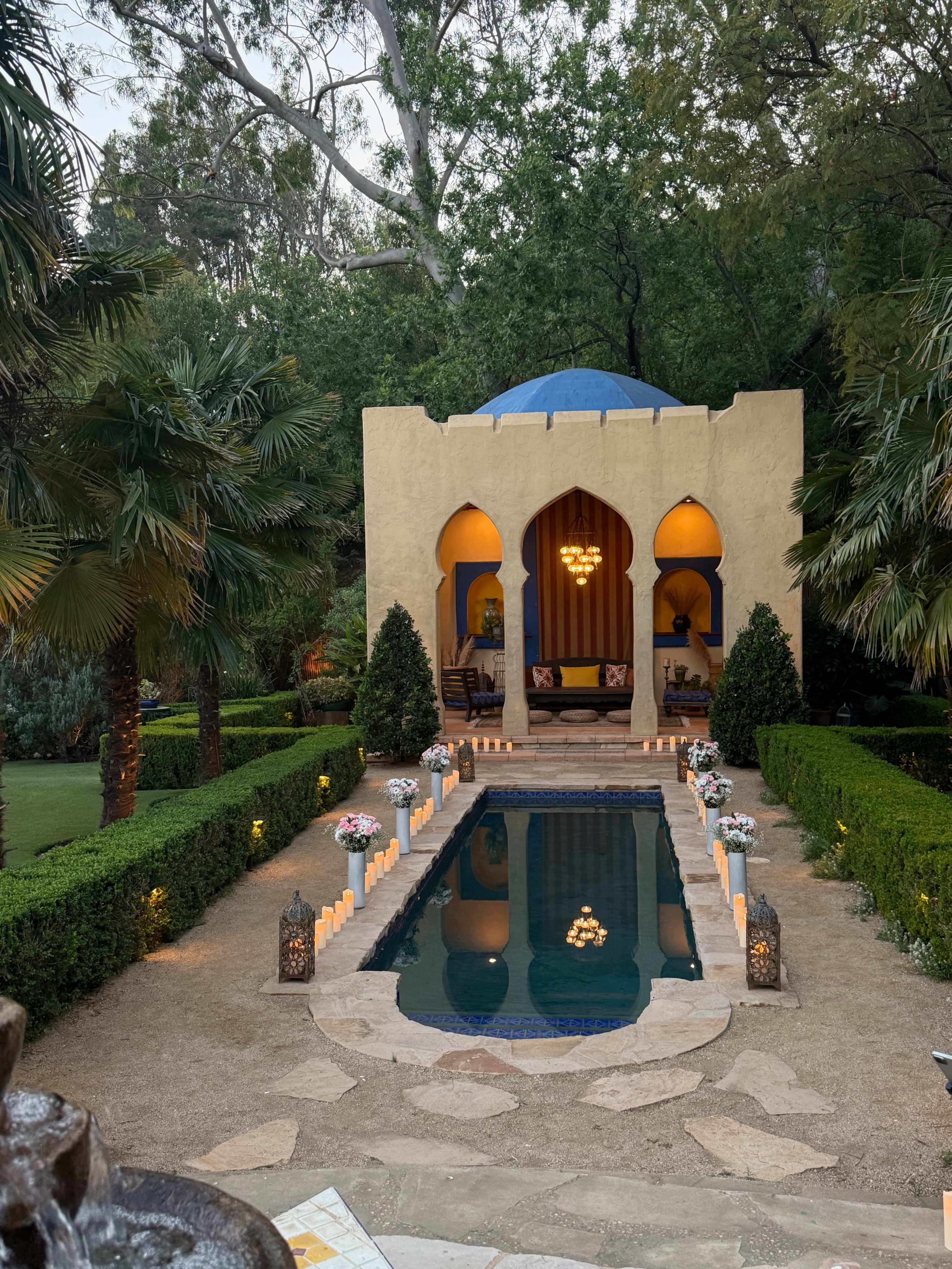 The image shows a tranquil garden featuring a small, arched pavilion with a blue dome, surrounded by neatly trimmed hedges and palm trees, and a reflecting pool lined with candles.