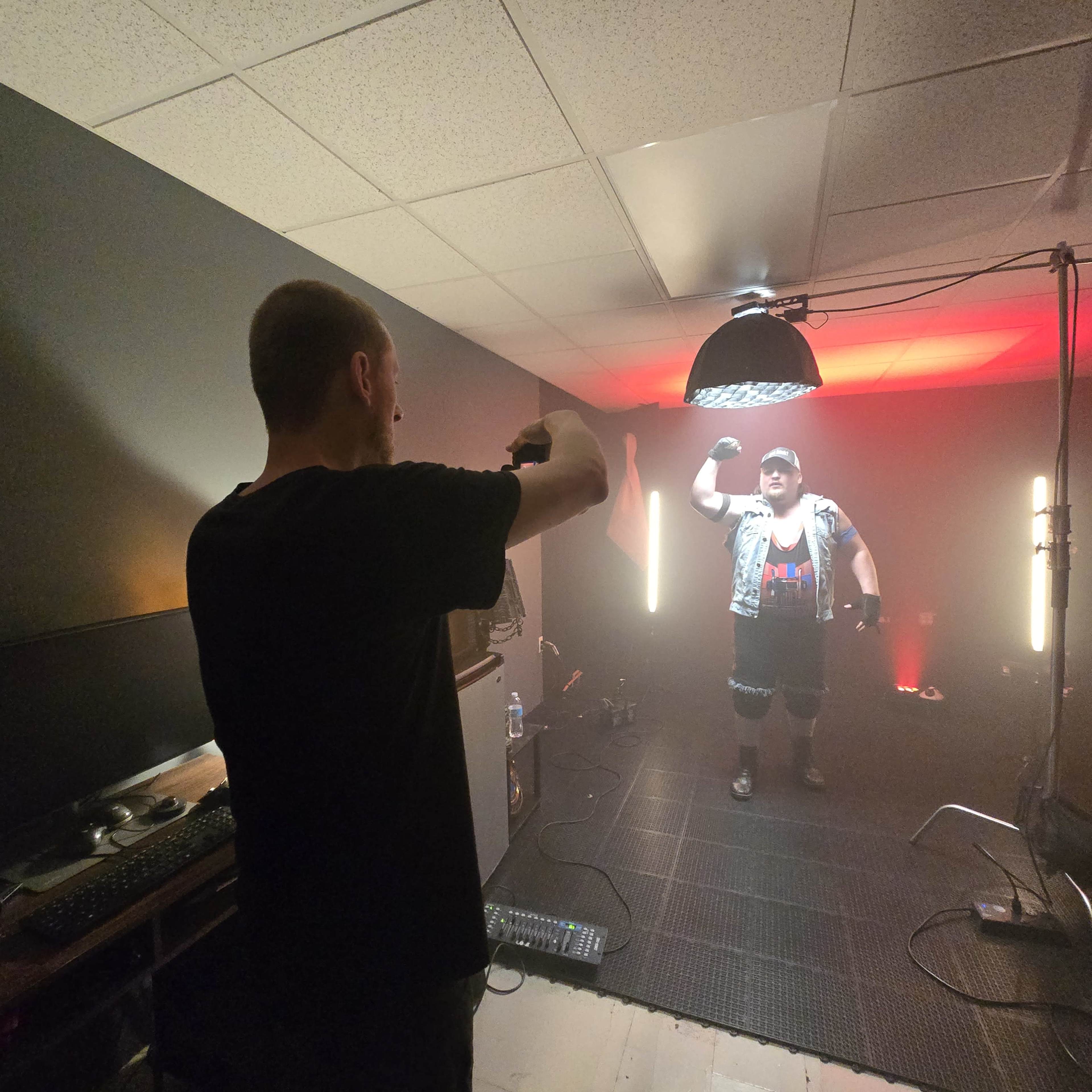 A person is taking a photograph of a muscular individual posing in a dimly lit room with colored lights and studio equipment.