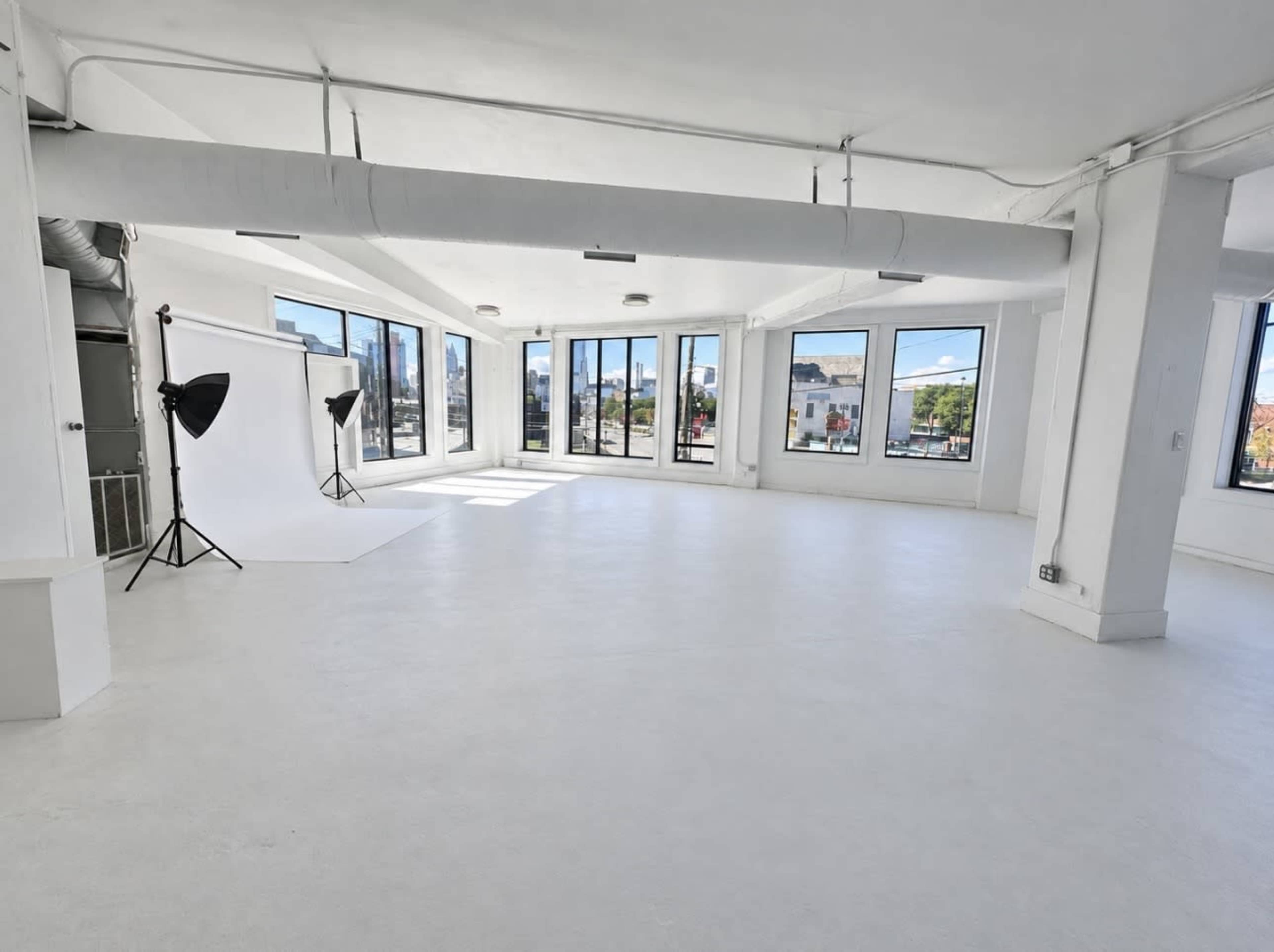 The image shows a spacious, bright photo studio with large windows, white walls, and a seamless backdrop setup in one corner.