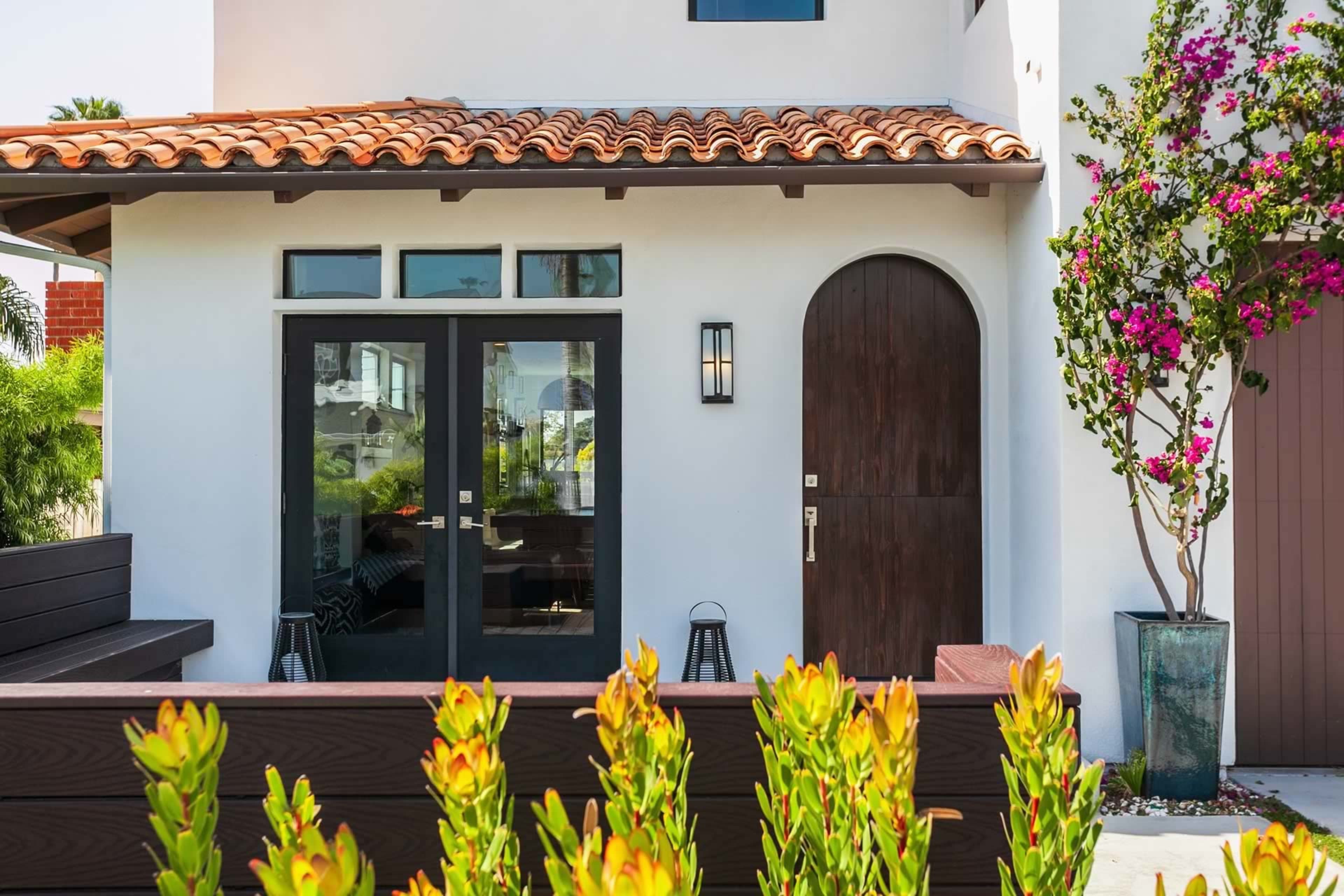A modern home with a red tile roof, dark wooden doors, and a surrounding garden featuring green plants and a flowering bush.