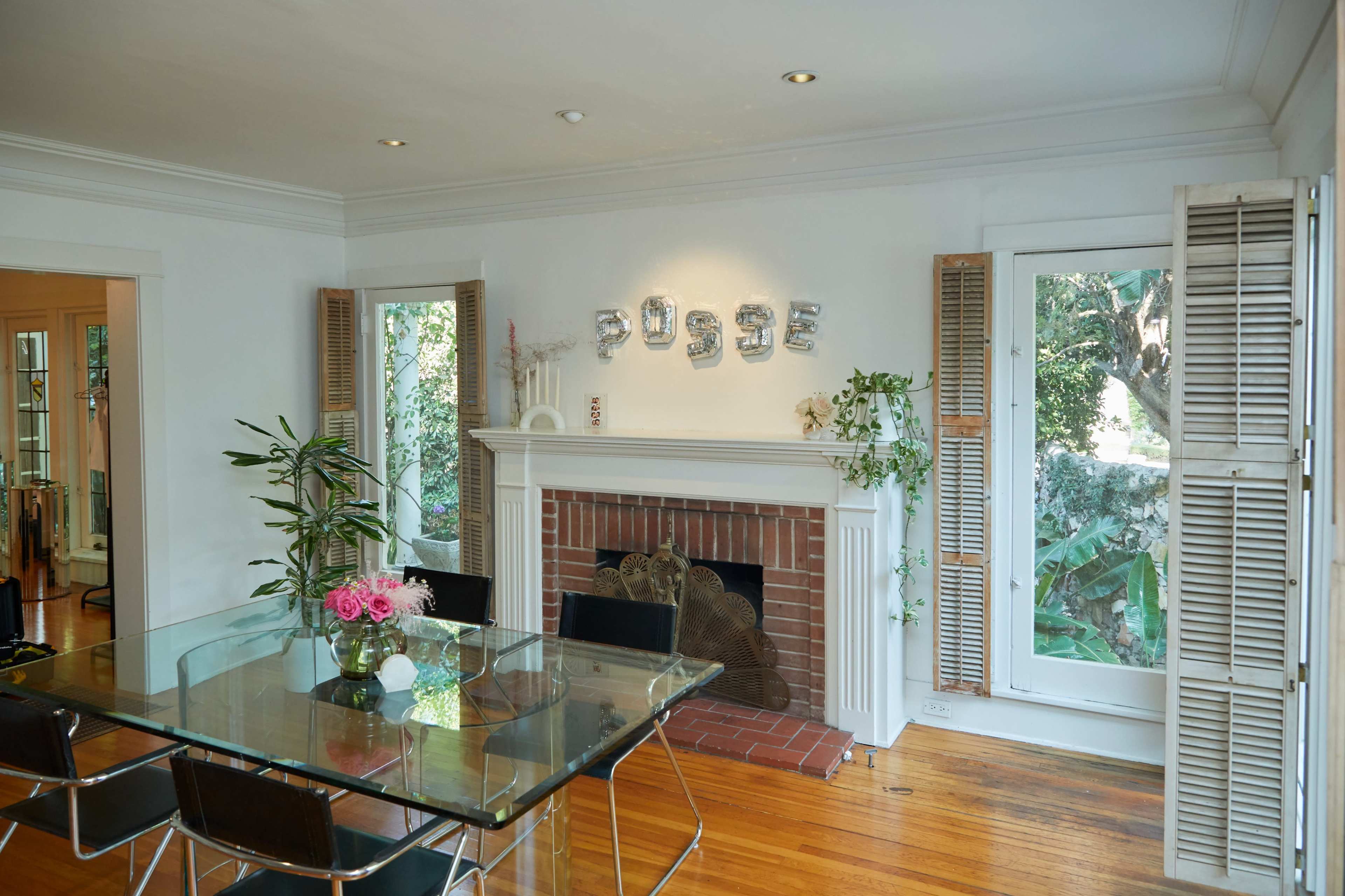 A dining area features a glass table surrounded by black chairs, with a fireplace and large windows that showcase greenery outside.