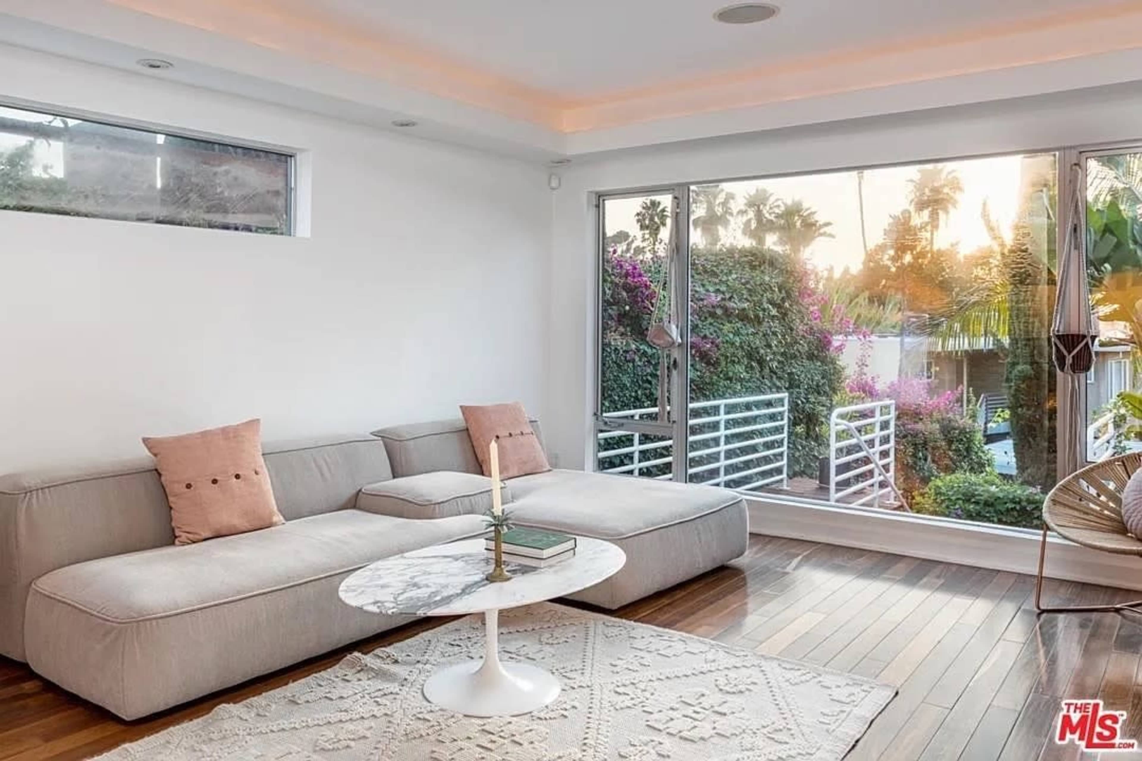 The image shows a living room featuring a light-colored sectional sofa, a round marble coffee table, and large glass doors leading to a garden area filled with greenery and flowers.