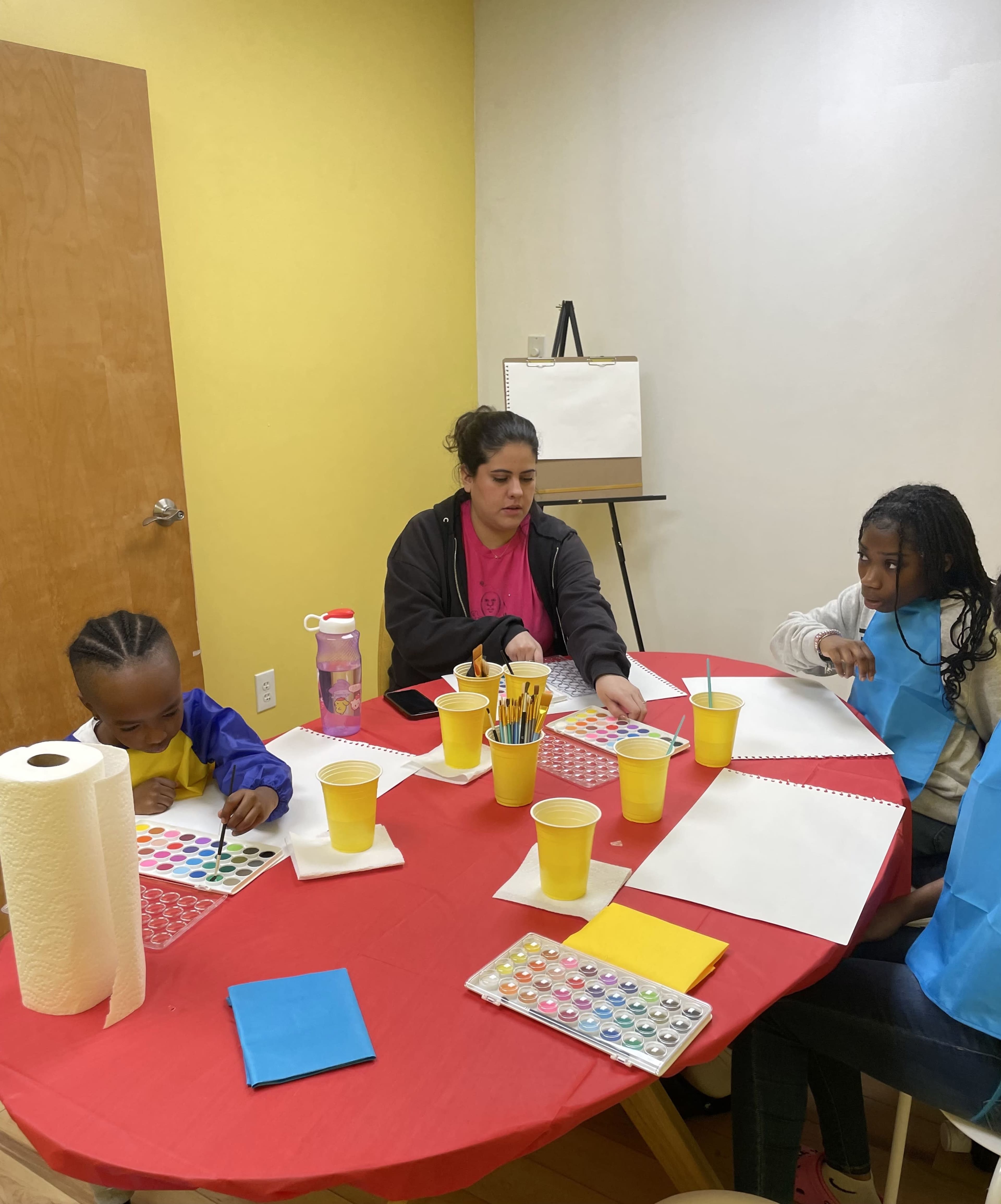 Three children and an adult are engaged in a painting activity at a table covered with art supplies, including yellow cups, watercolor palettes, and blank sheets of paper.