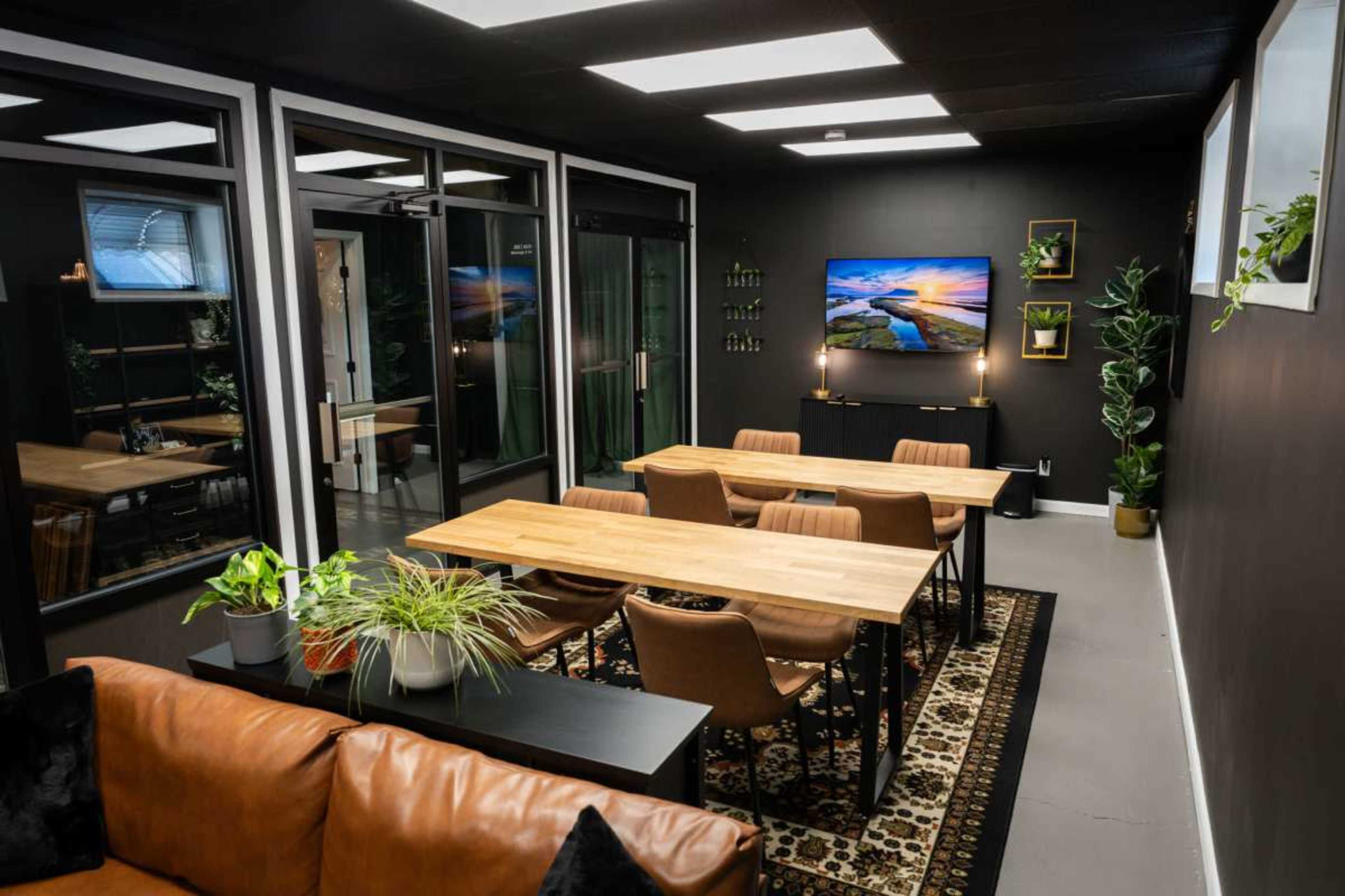 The image shows a modern office meeting room with wooden tables and chairs, a large television on the wall, and plants positioned around the space.