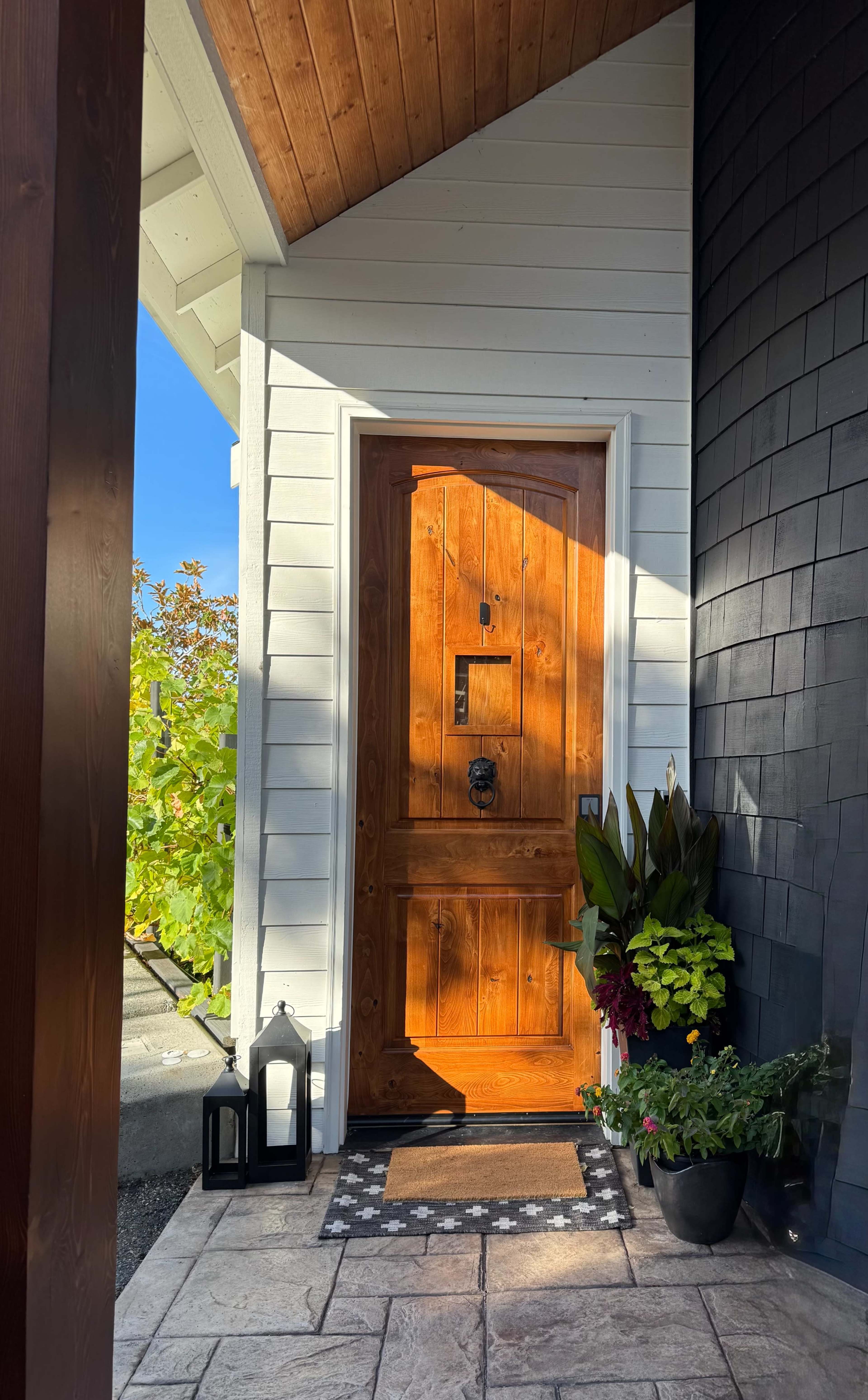The image shows a wooden front door with a diagonal design, framed by plants and a decorative mat on a stone pathway under a sunny sky.