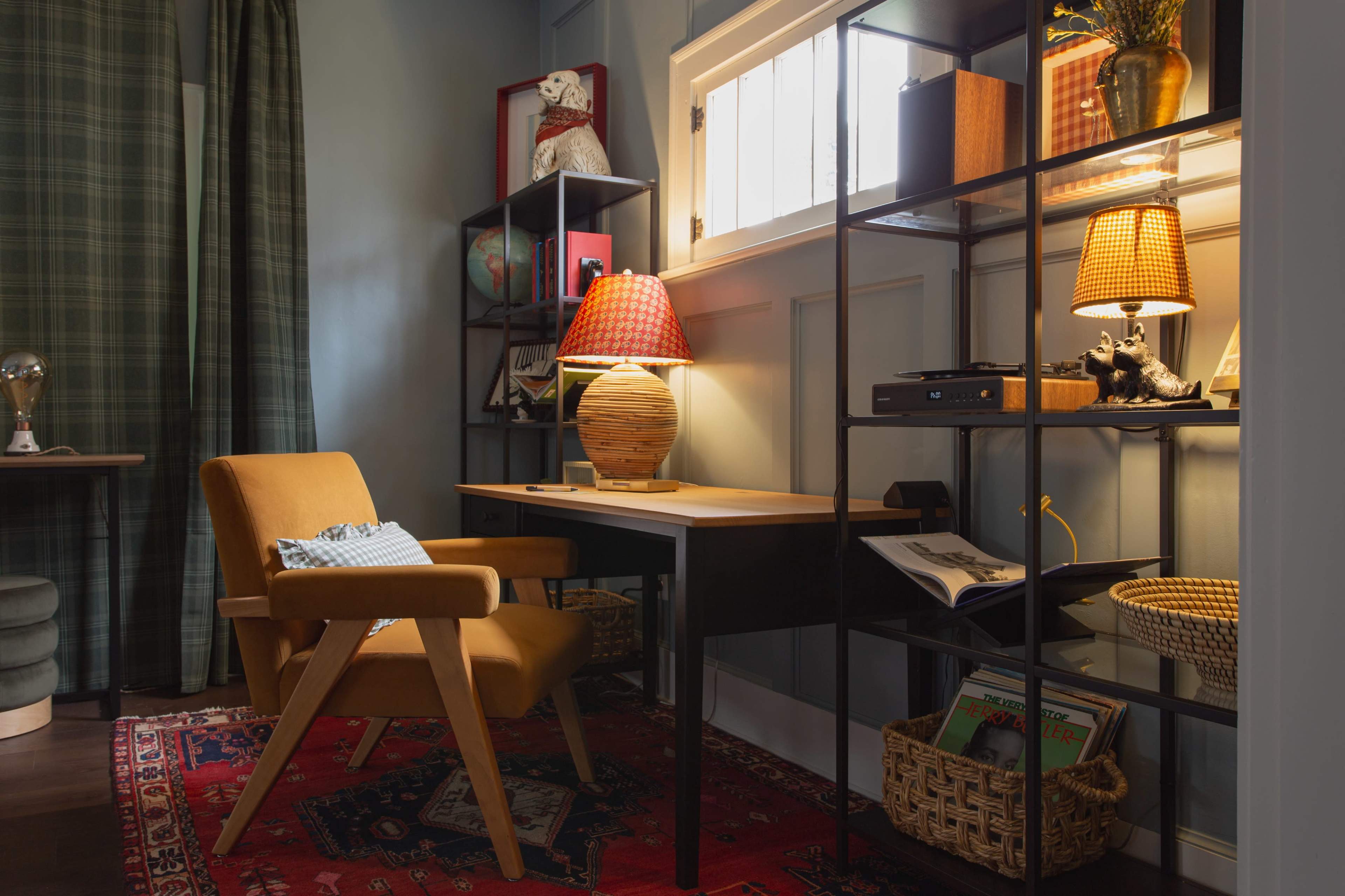 A cozy home office features a wooden desk, a comfortable orange armchair, and two lamps illuminating a shelf filled with books and decorative items.