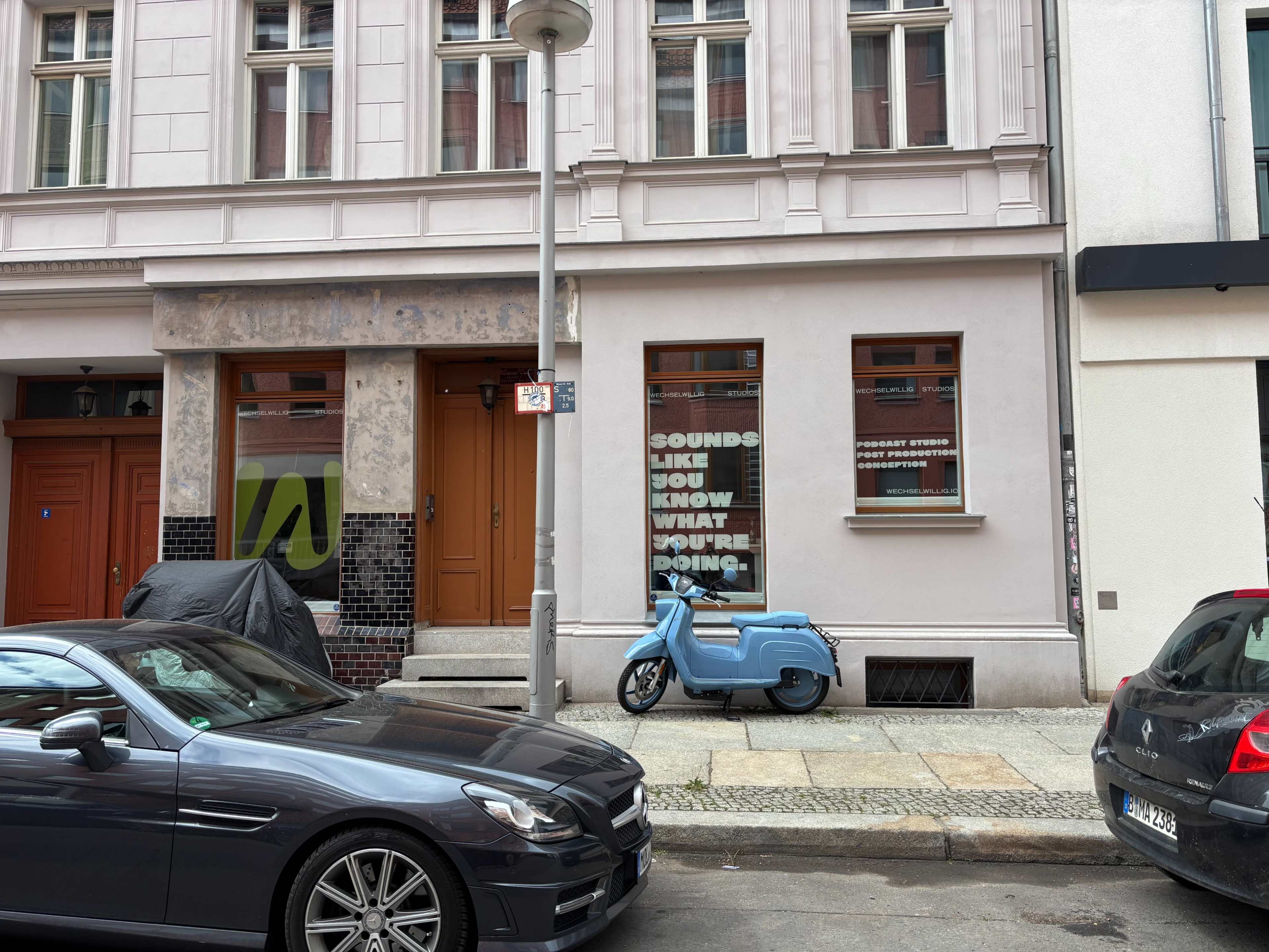 A light blue scooter is parked on the street in front of a building with large windows displaying the text "SOUNDS LIKE YOU KNOW WHAT YOU'RE DOING."