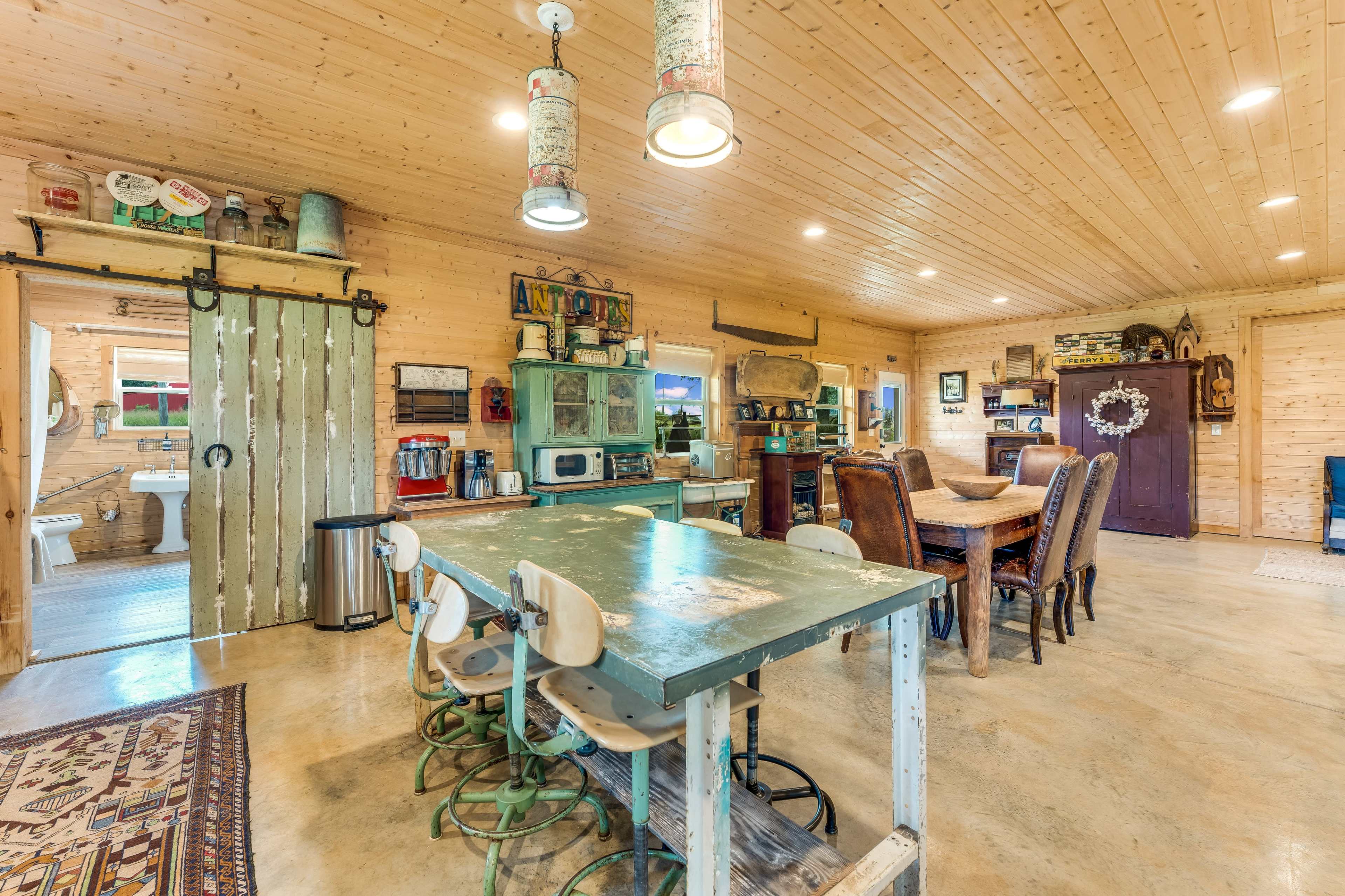 The image shows a spacious wooden interior of a rustic dining area with a large table, several chairs, and a visible kitchen area in the background.