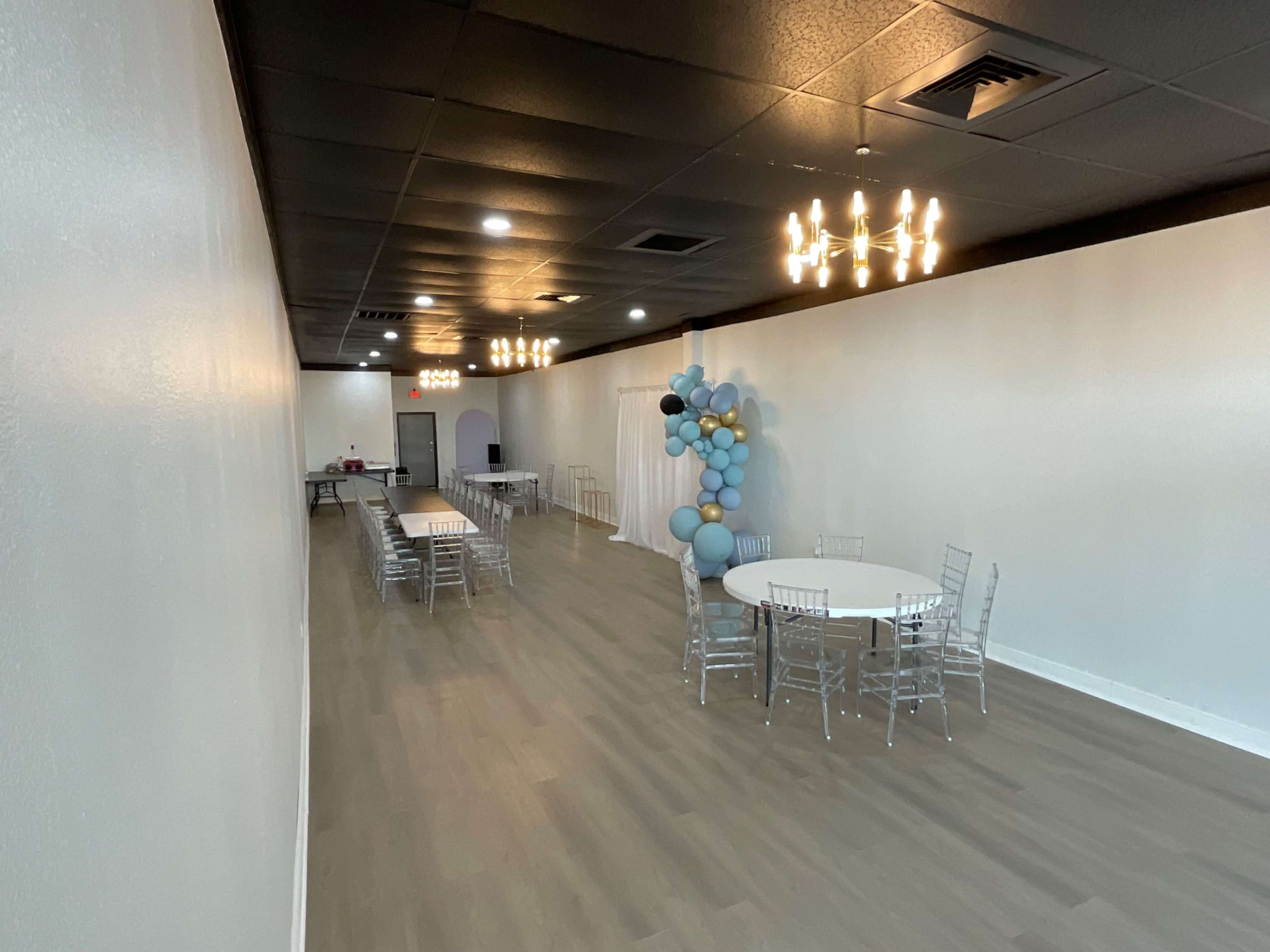 The spacious interior features several round tables with clear chairs, illuminated by modern light fixtures, and a balloon display against one wall.