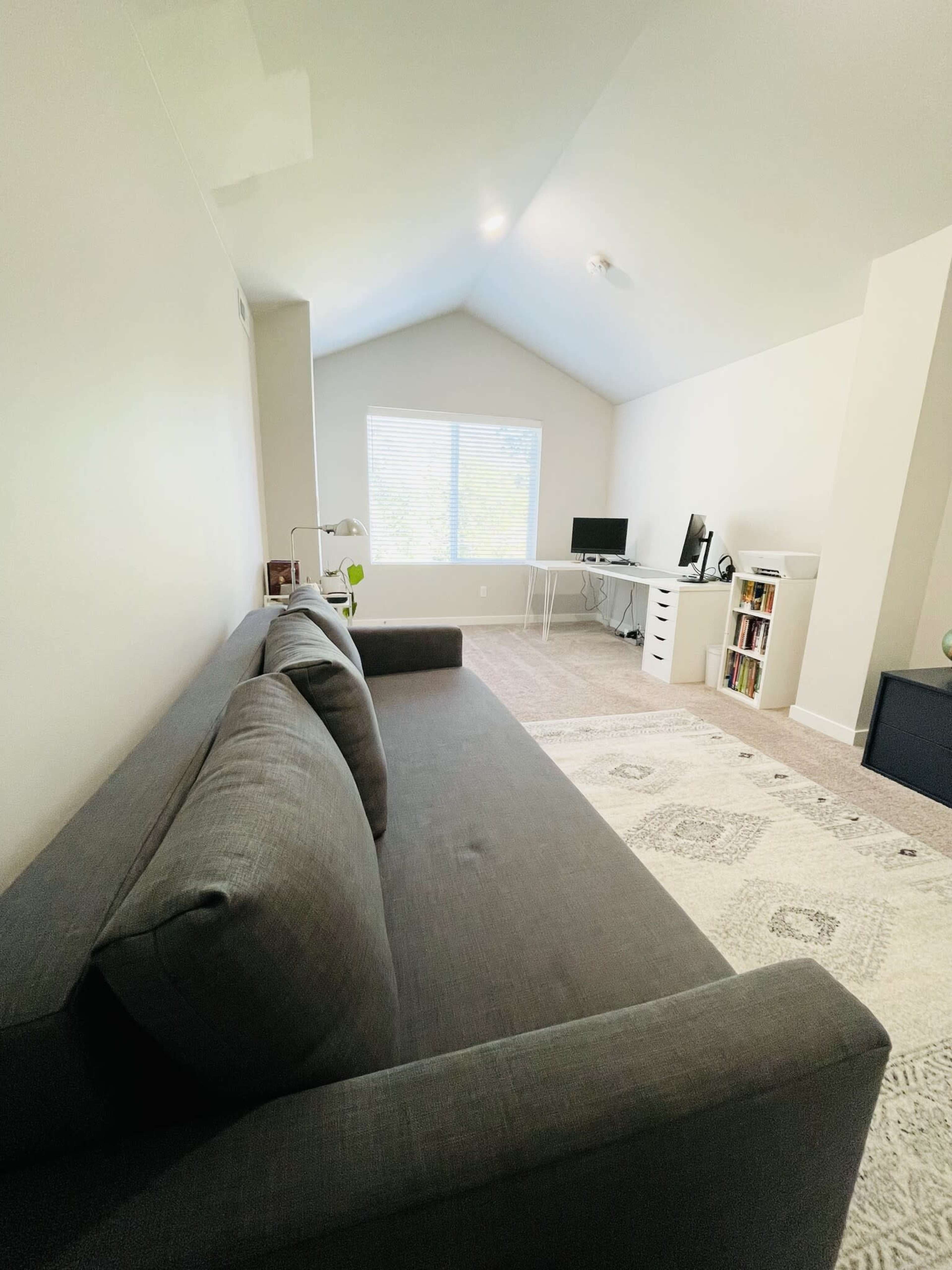A gray sofa is positioned in a room with a peaked ceiling, a window providing natural light, and a work desk with a computer.