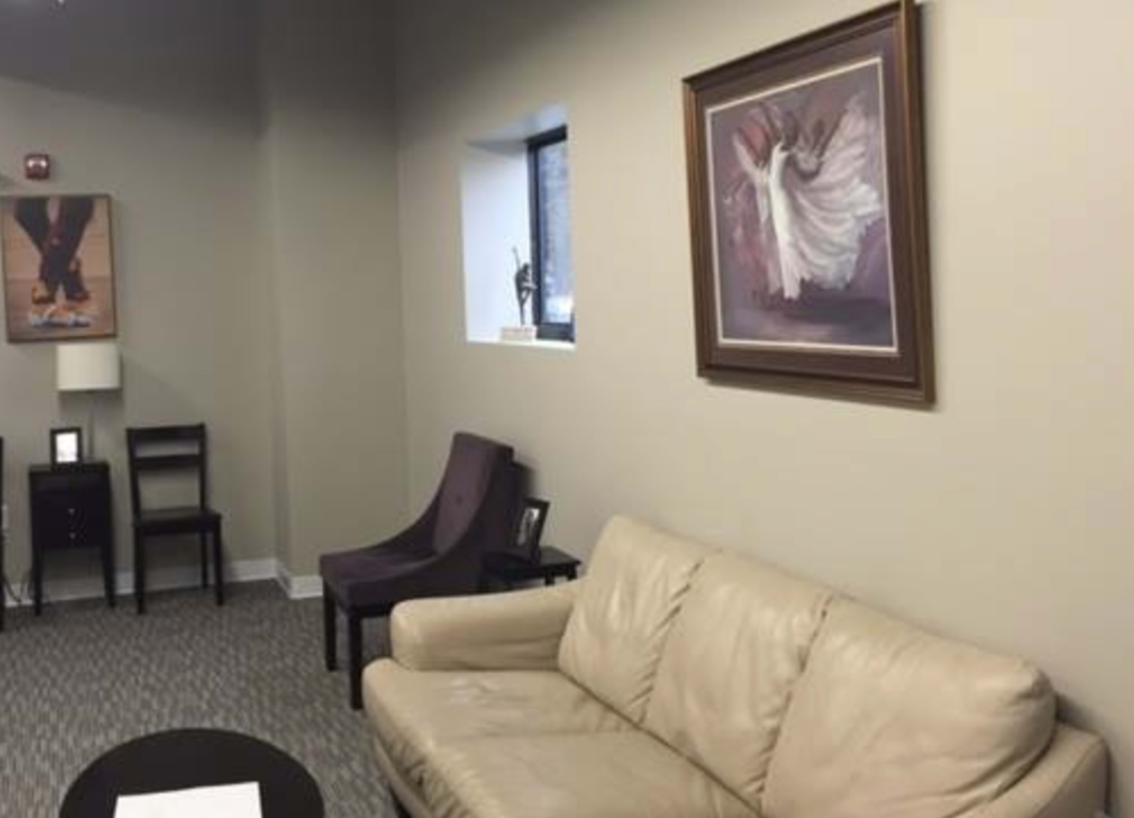 The image shows a modern waiting room with a light-colored sofa, a dark table, a chair, and a wall adorned with framed artwork.