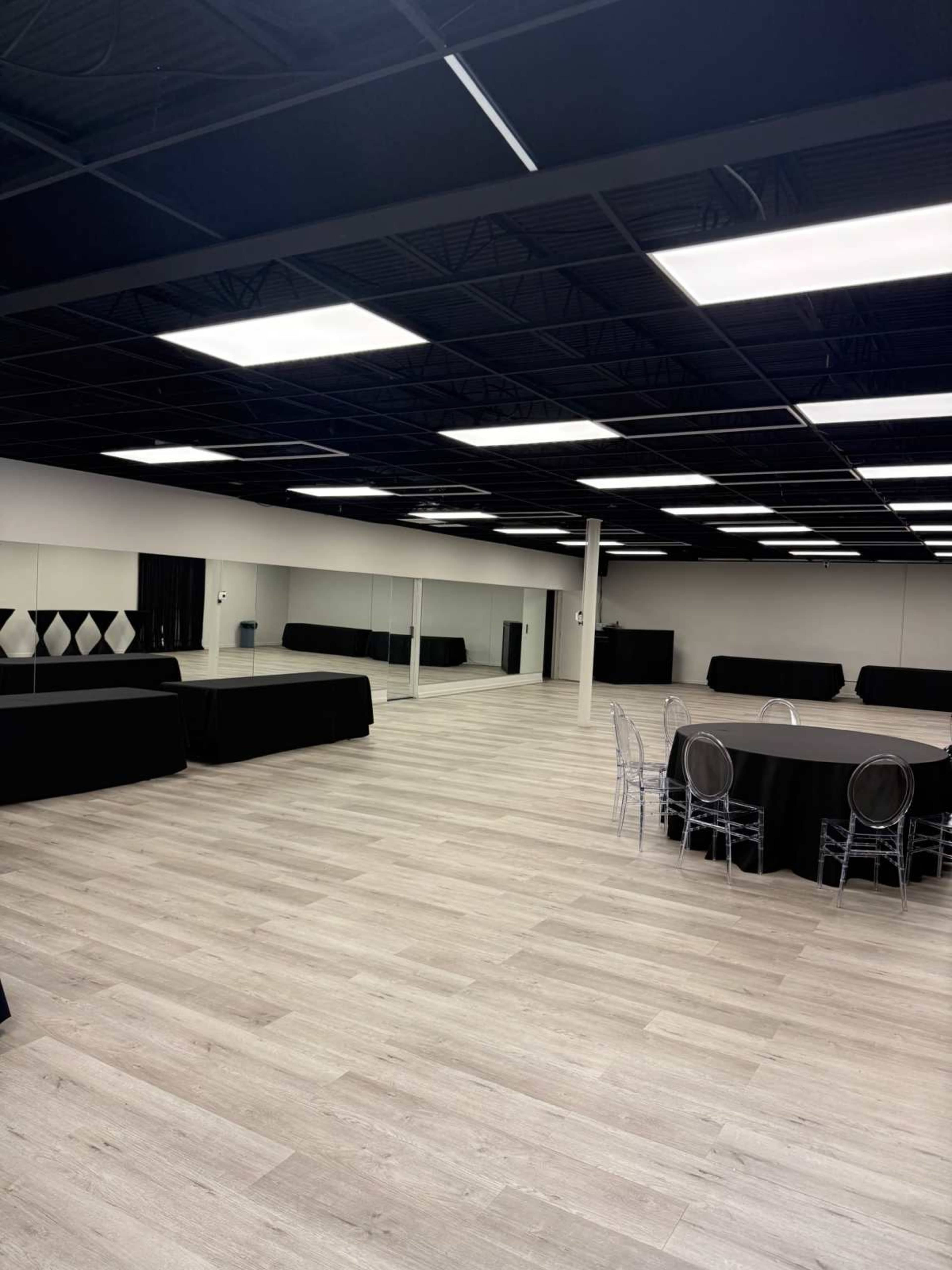 The image shows a spacious, well-lit event room featuring light-colored wooden flooring, multiple tables covered with black cloth, and a mirrored wall.