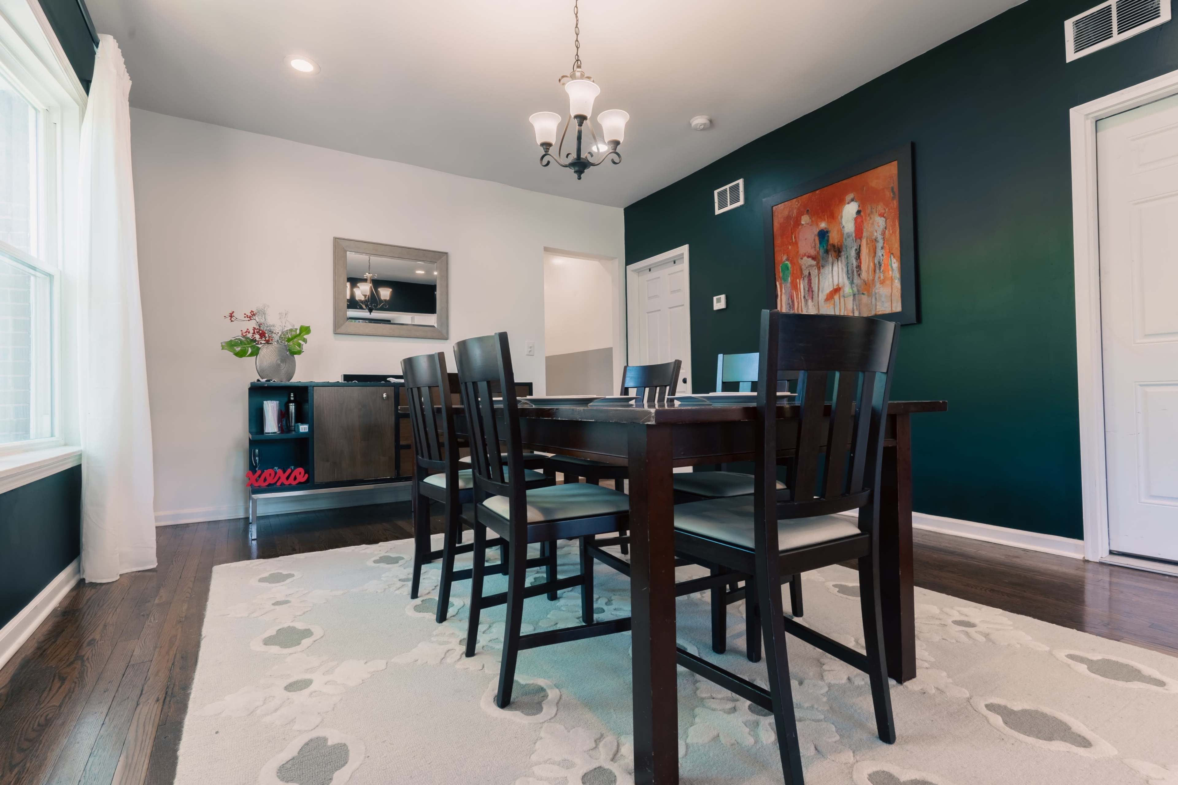 A dining room features a dark wall, a wooden table surrounded by black chairs, and a chandelier overhead, with a cabinet and artwork on the walls.