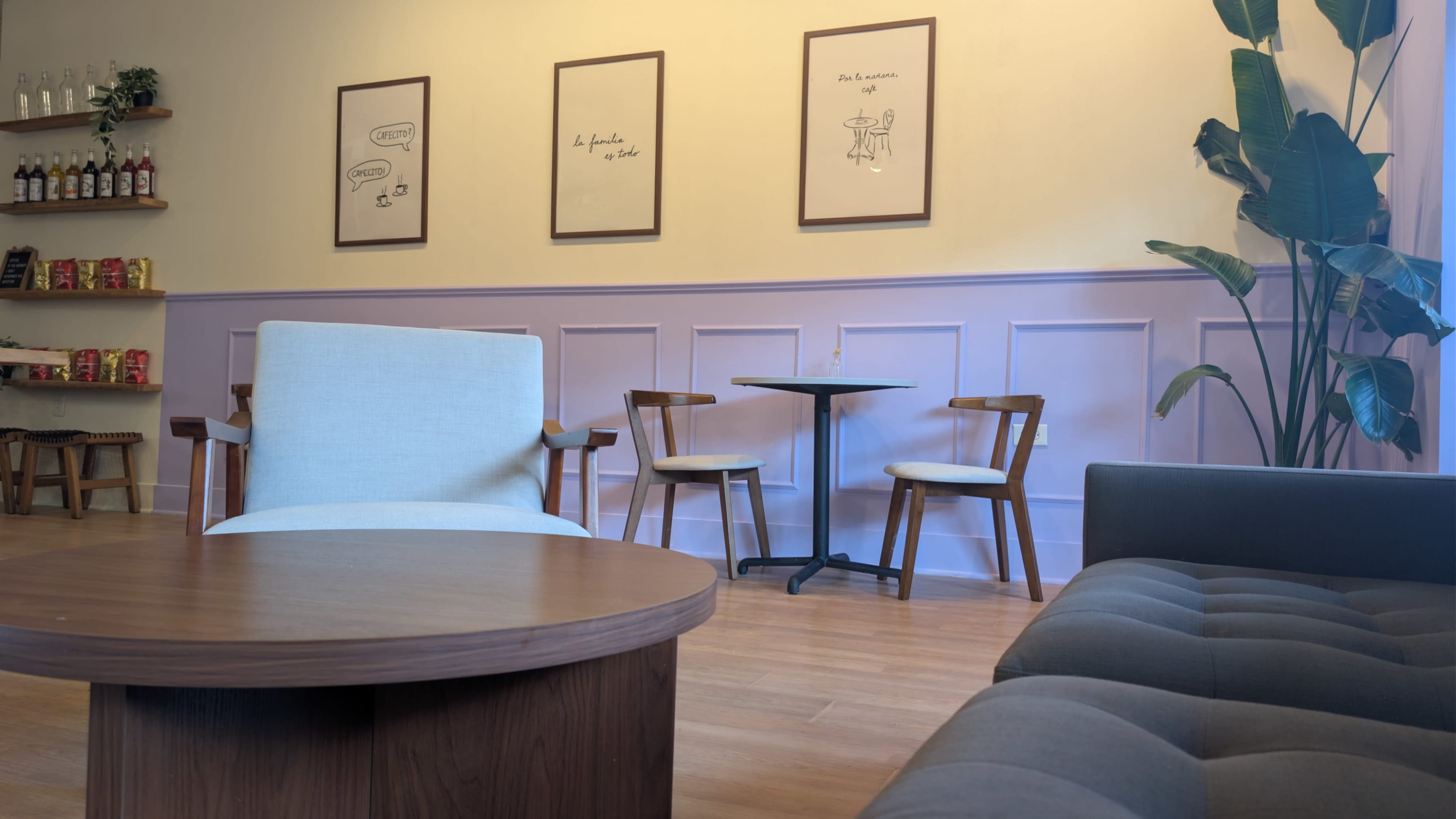 The image shows a cozy cafe interior with a light-colored wall, framed artwork, and a small table with two chairs, along with a beige armchair and a dark sofa.