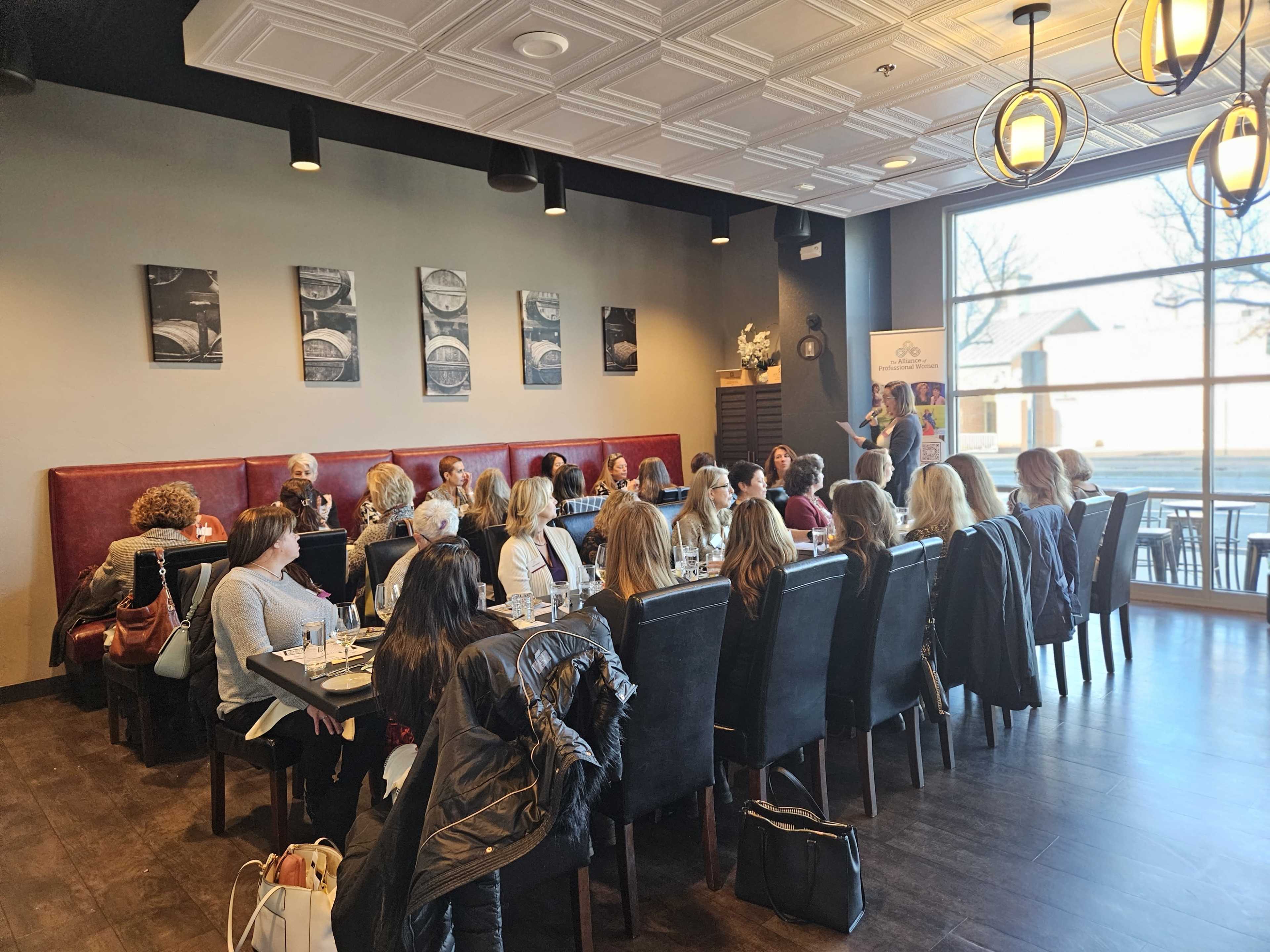 A group of women is seated at a long table in a restaurant, engaged in conversation while a speaker addresses them from the front.