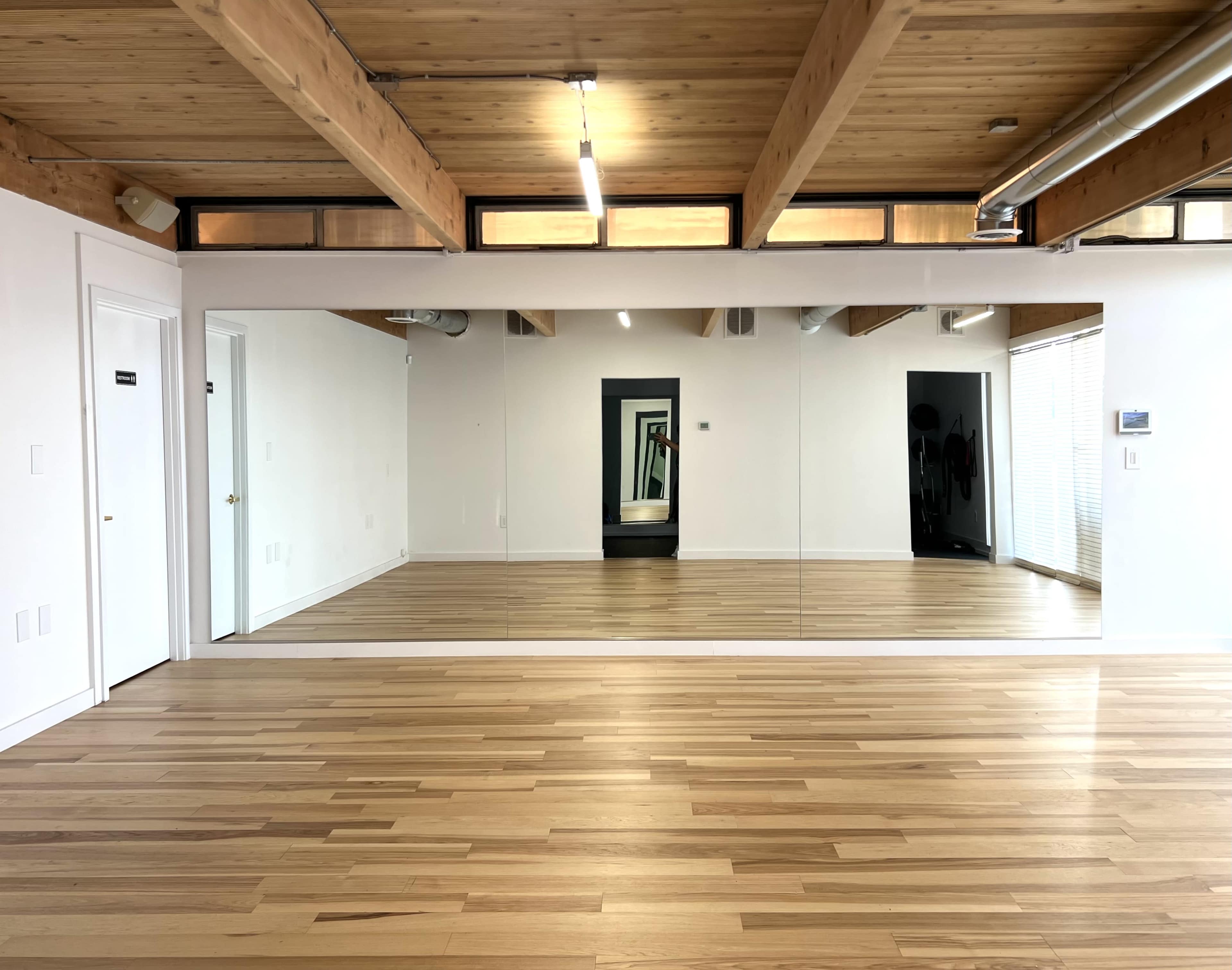A bright room with wooden beams and flooring, featuring a large mirror along one wall.