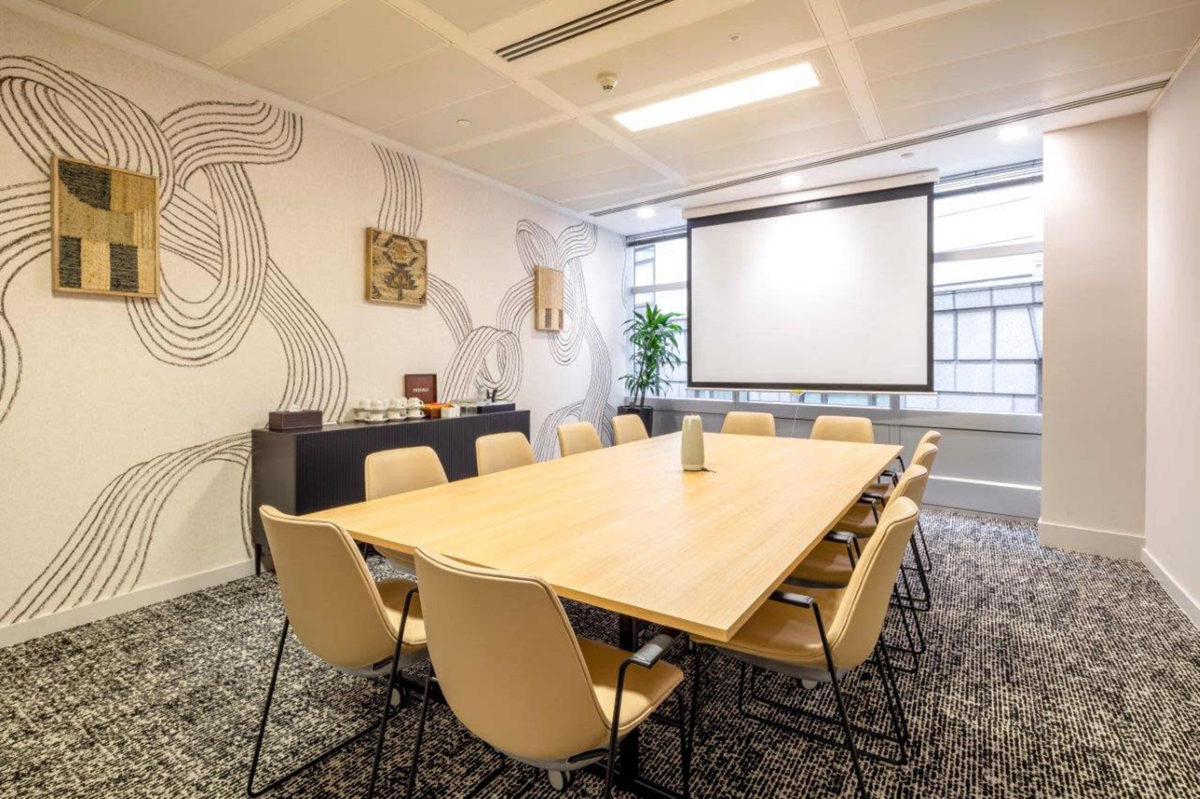 A conference room is set with a large wooden table surrounded by beige chairs, featuring a projector screen and decorative wall art.