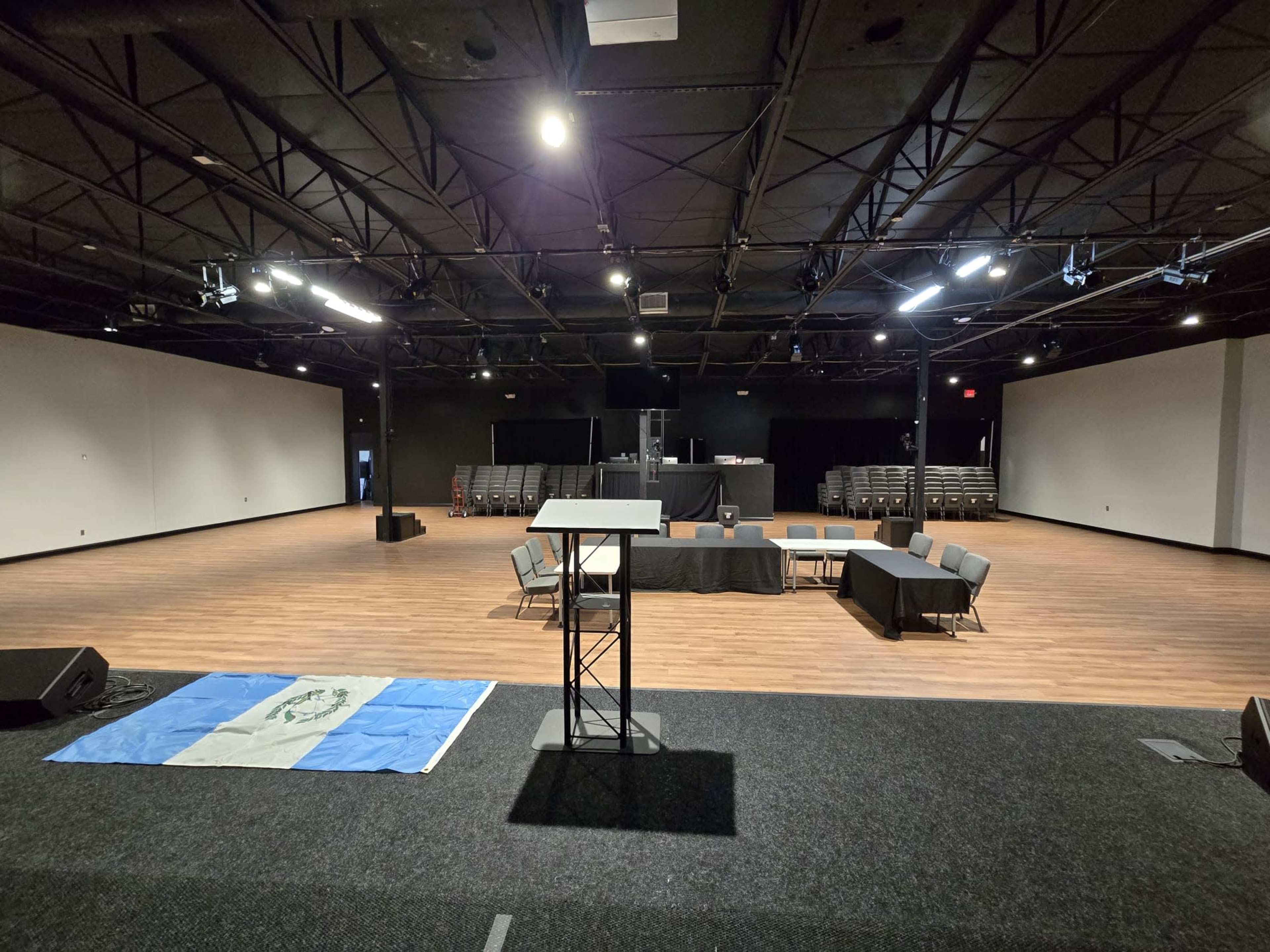 The image shows a large, empty event space with wooden flooring, a podium, chairs arranged around a central table, and a flag on the floor.