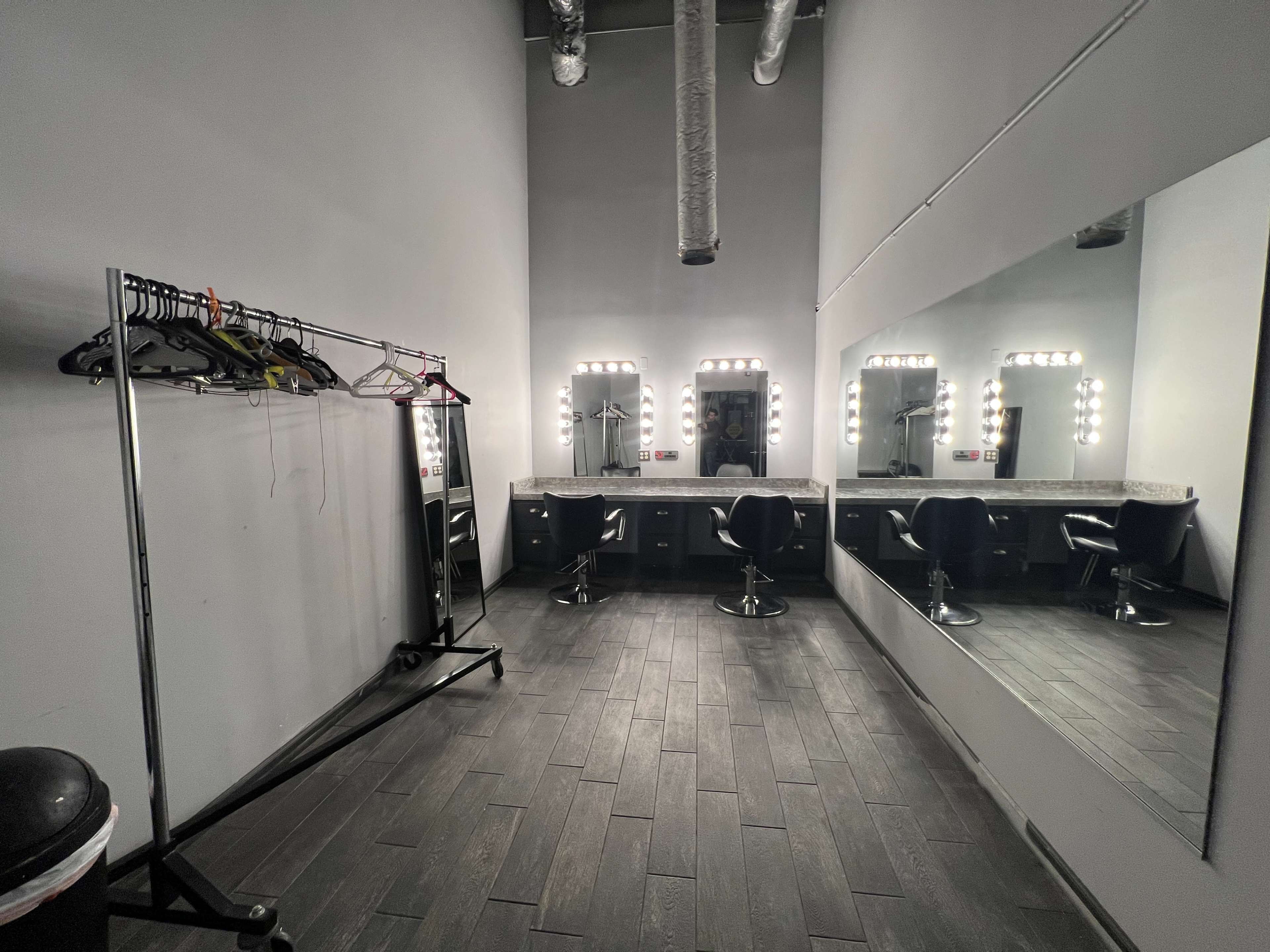 The image shows a makeup and dressing room with a mirrored wall, bright vanity lights, a clothing rack, and black chairs.