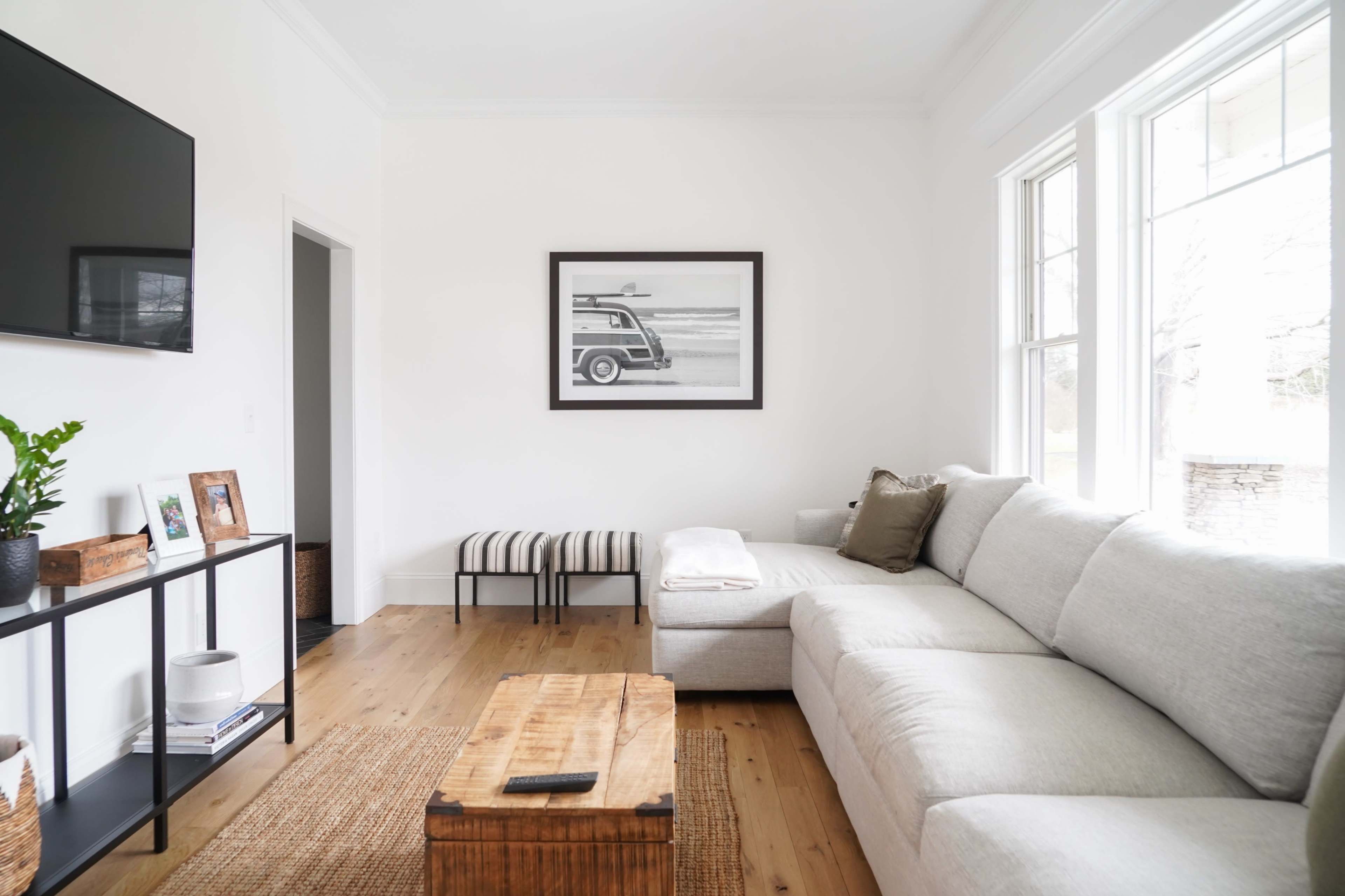 A light-colored living room features a sectional couch, a wooden coffee table, a striped bench, and a wall-mounted television.