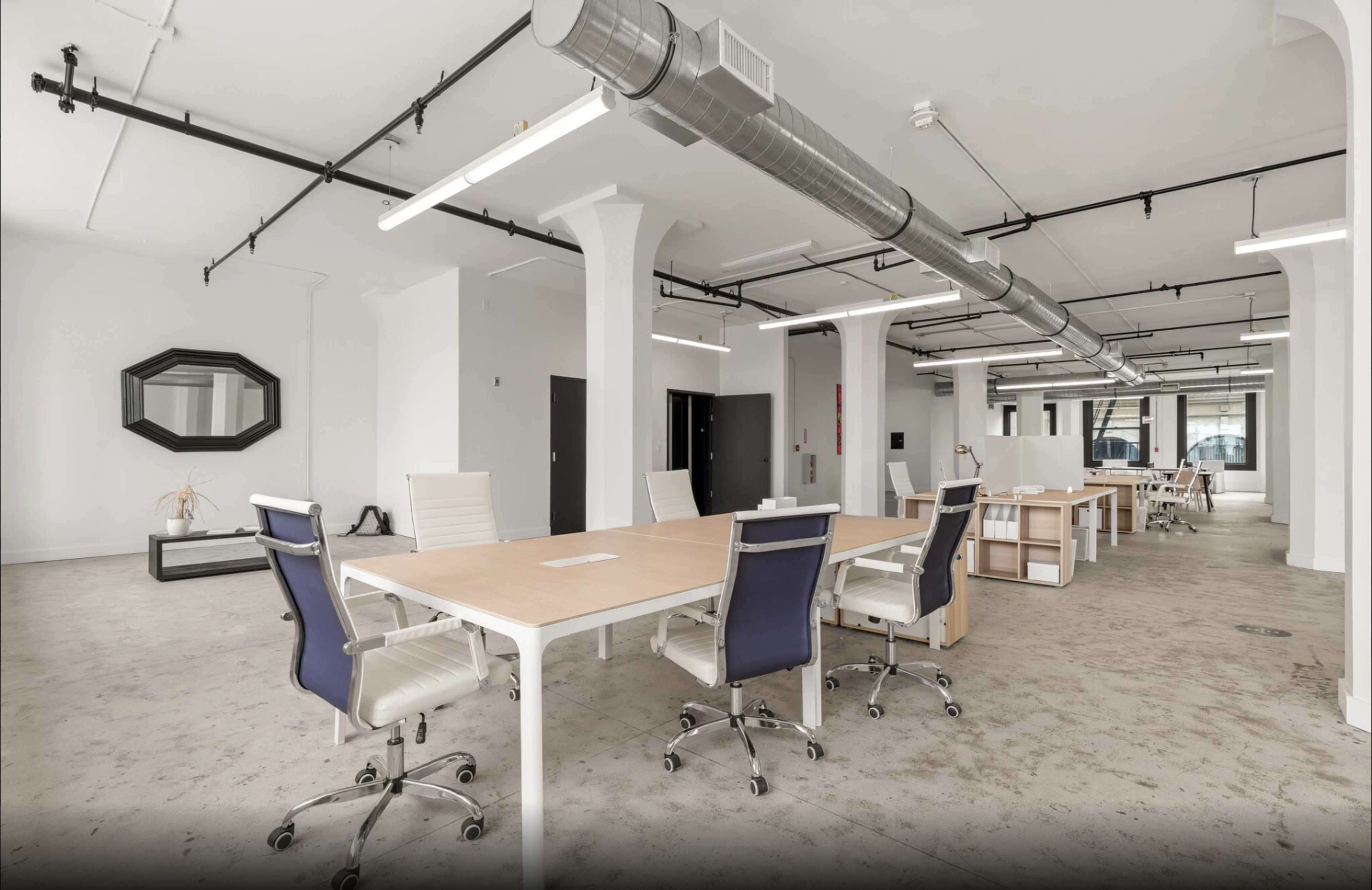 The image shows a spacious, modern office interior with multiple workstations arranged around a large central table, featuring a polished concrete floor and ample overhead lighting.