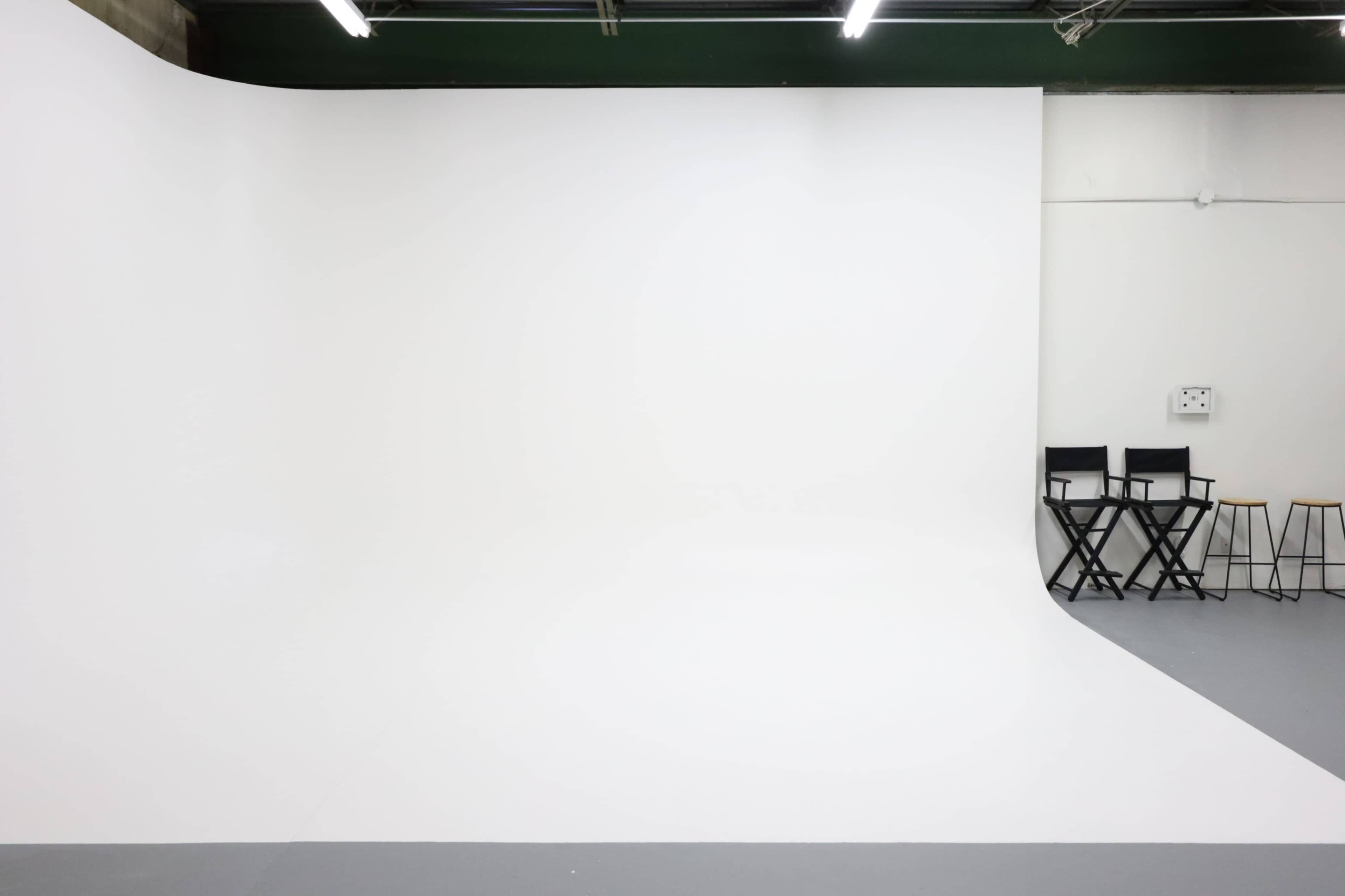 A minimalistic photography studio with a white backdrop and a few black chairs against a plain wall.