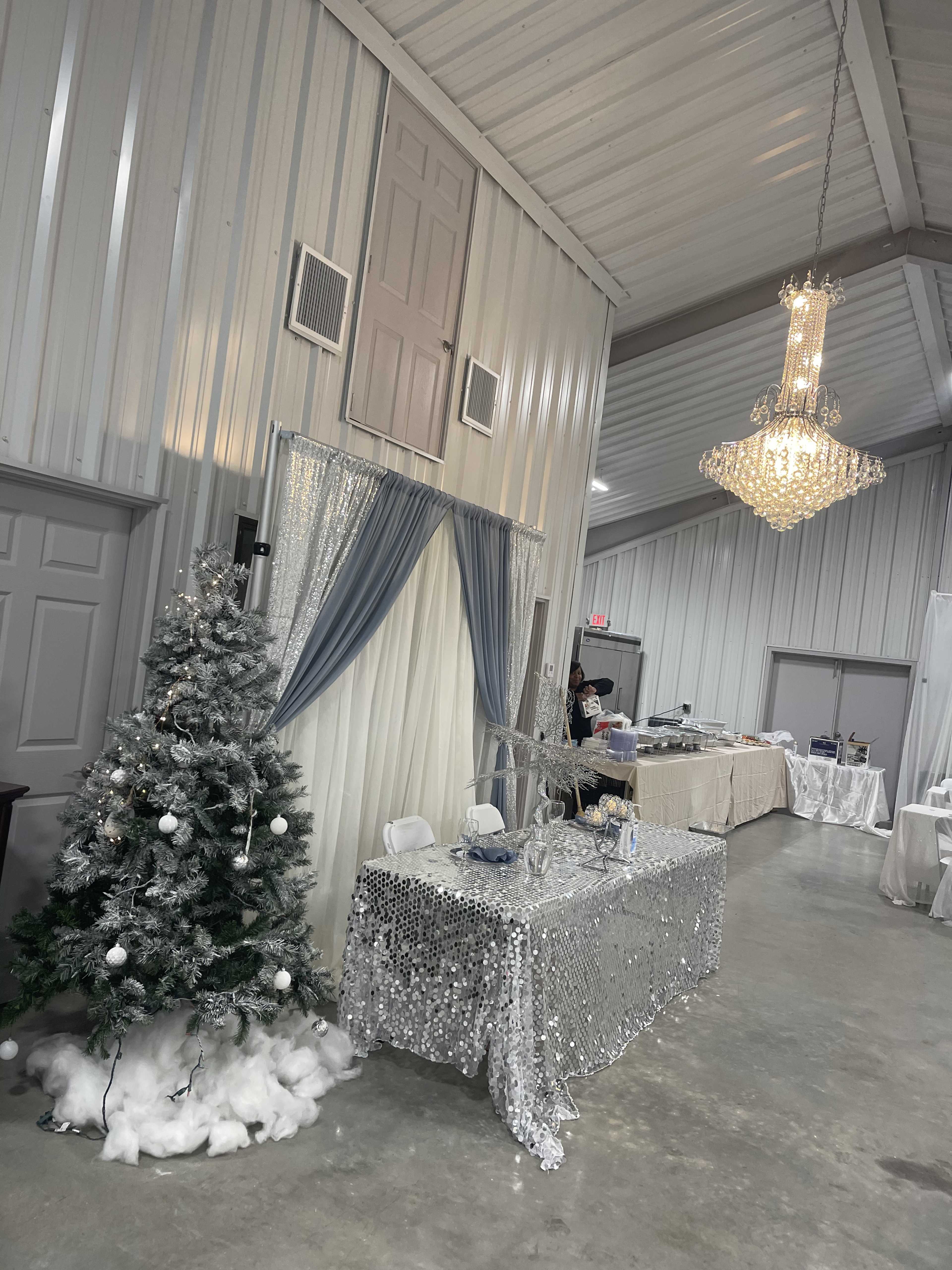A decorated event space with a silver tablecloth, a Christmas tree, and a chandelier, along with various tables set up in the background.