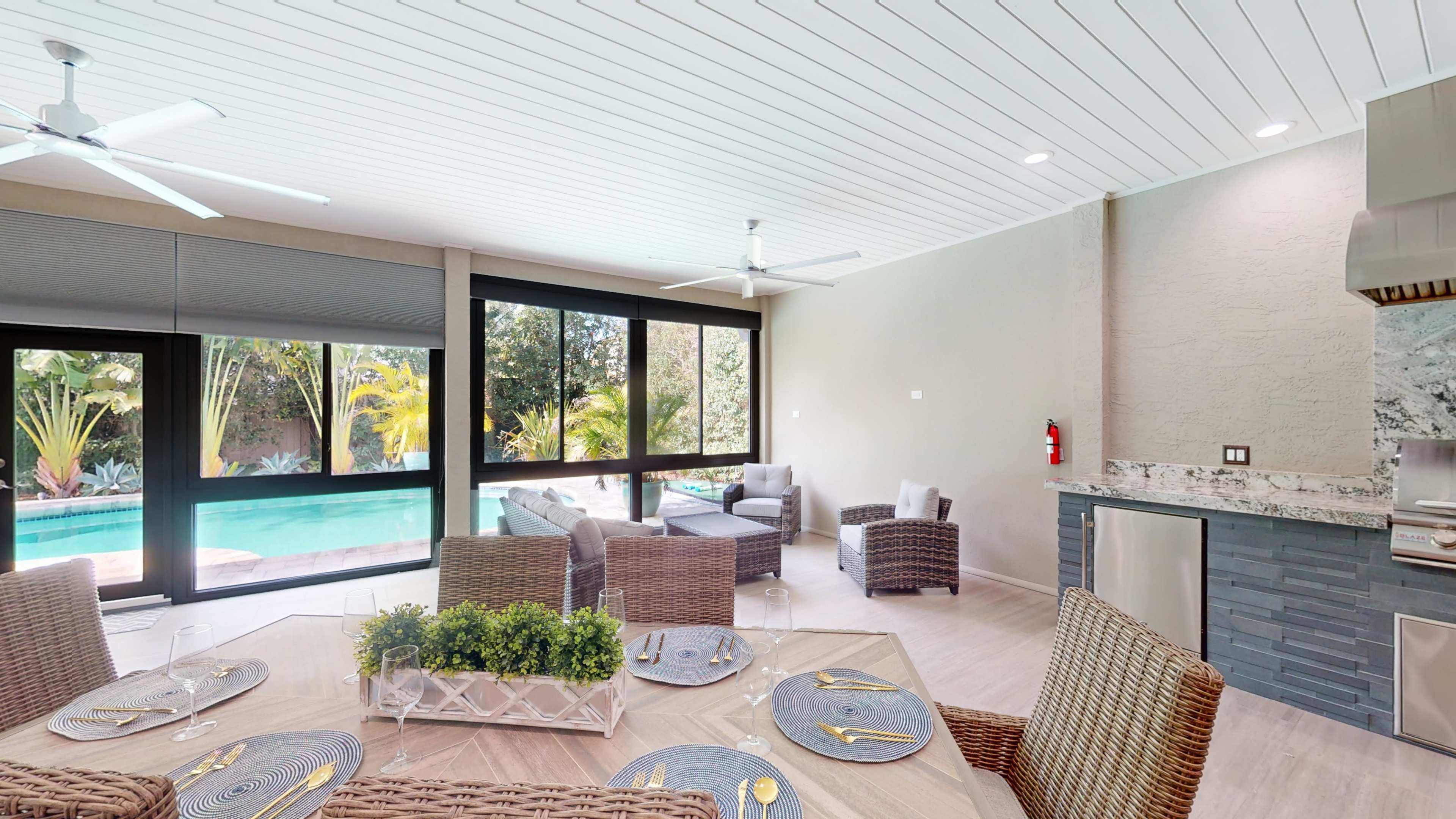 A modern open-concept dining area features a table set for four, surrounded by woven chairs, with large windows revealing a poolside view and lush greenery.