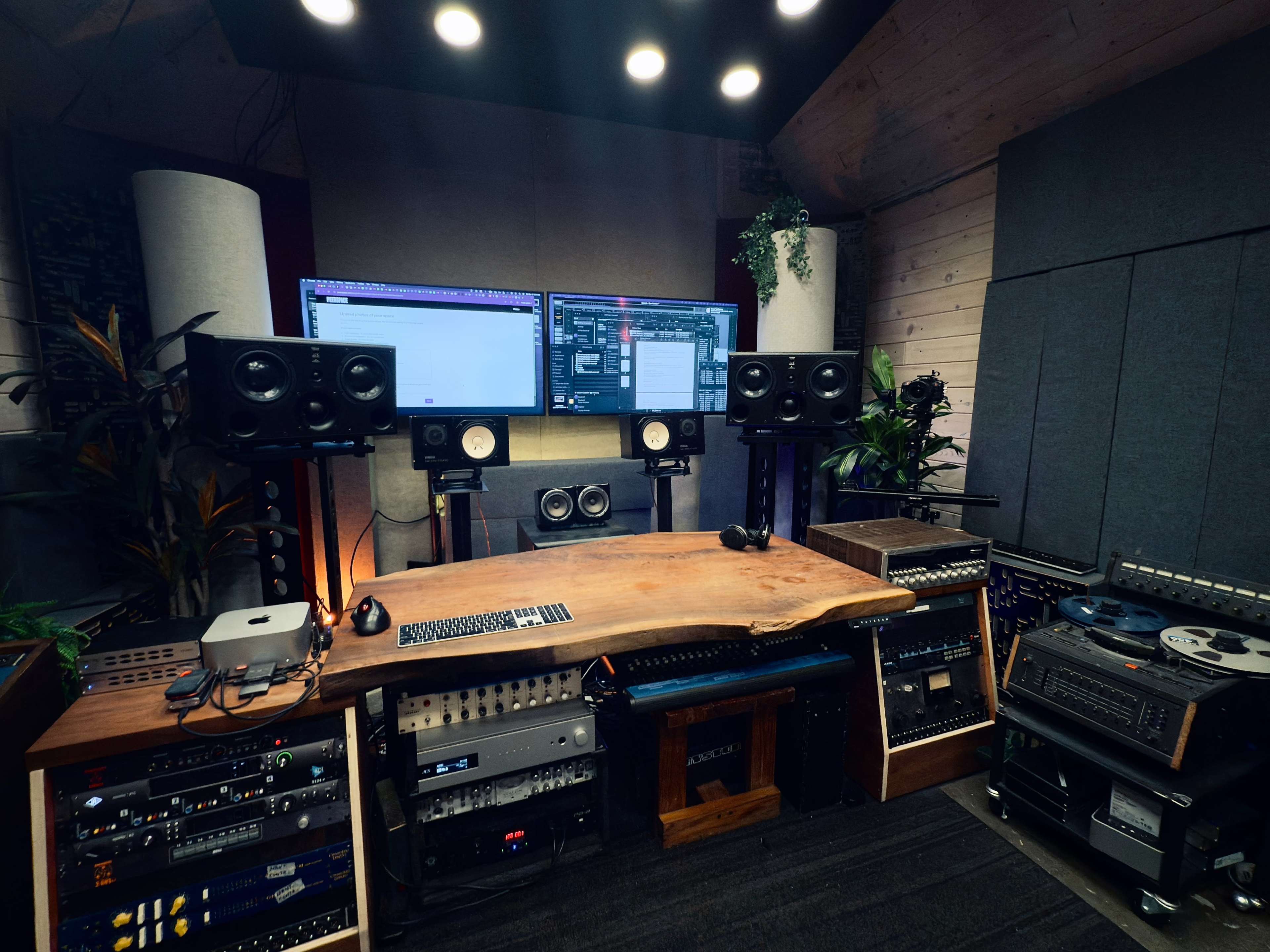 The image shows a music studio with two large monitors, multiple speakers, a wooden desk, and various audio equipment arranged neatly around the room.
