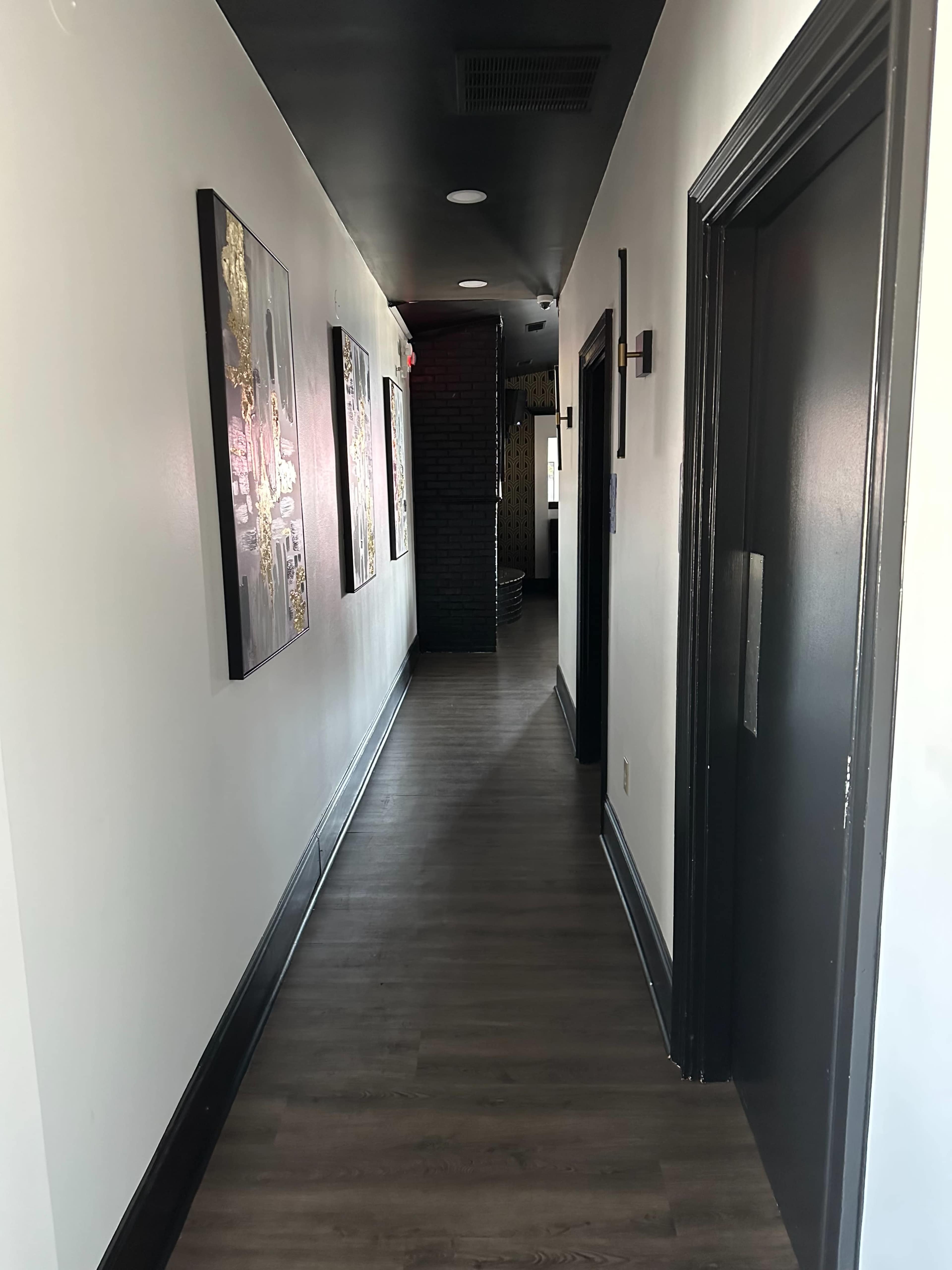 A narrow hallway with dark walls and wooden flooring is lined with framed artwork, leading towards an open doorway at the far end.