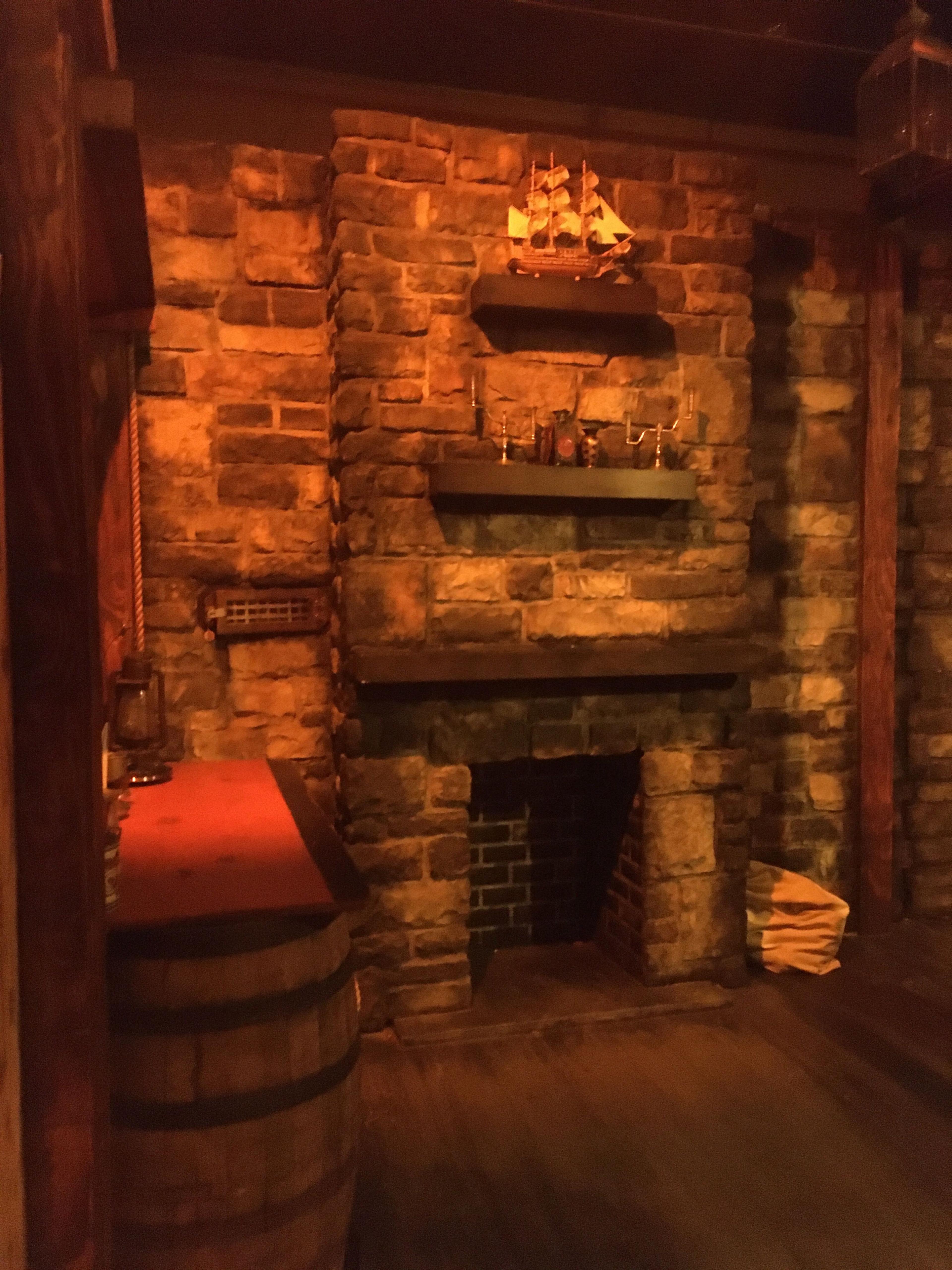A stone-walled room featuring a fireplace with a small ship model on a shelf above it, along with a wooden barrel and stacked papers nearby.