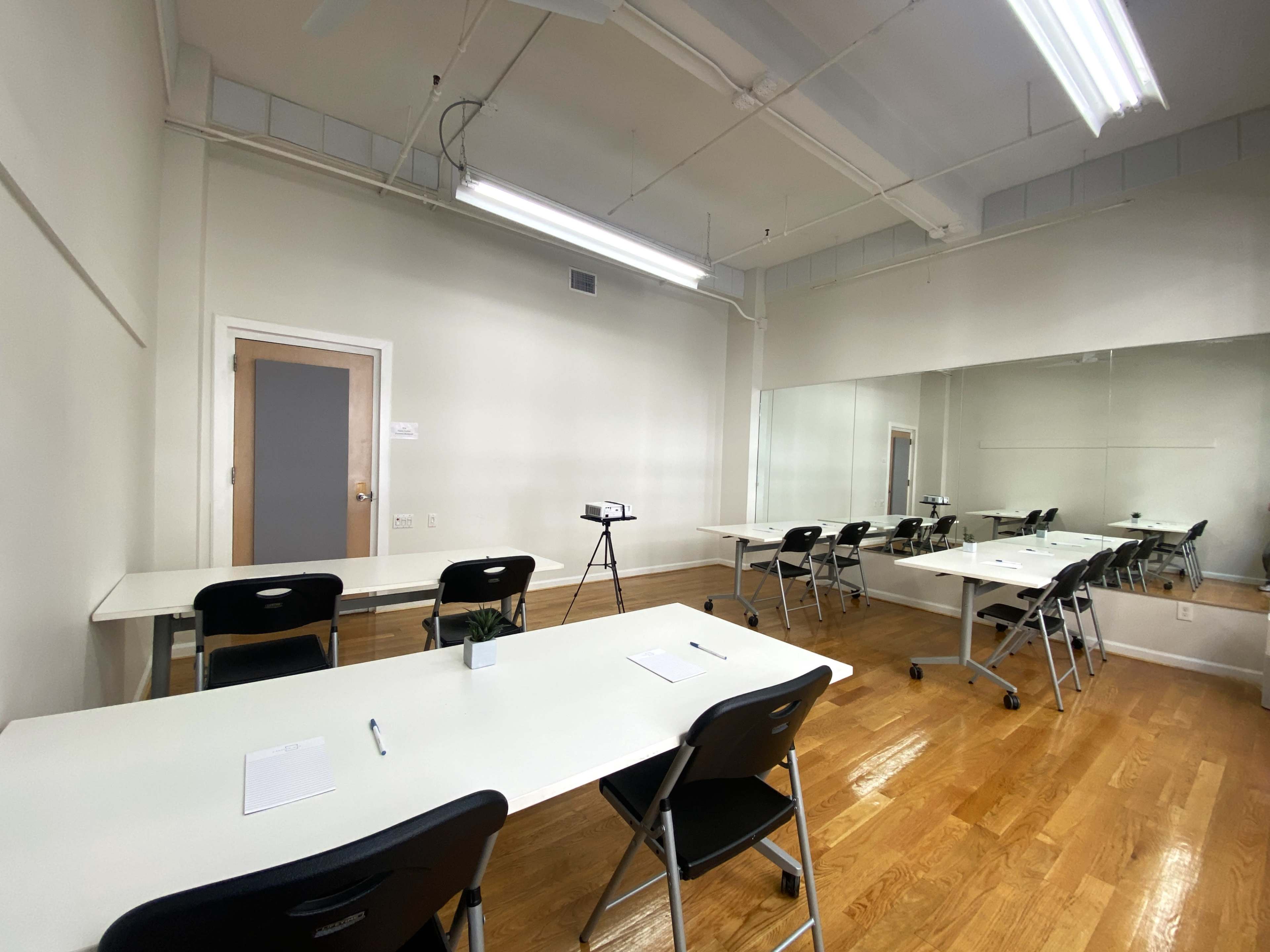 A minimalist room with several tables and chairs arranged for a meeting, featuring a large mirror along one wall and a tripod set up in front.