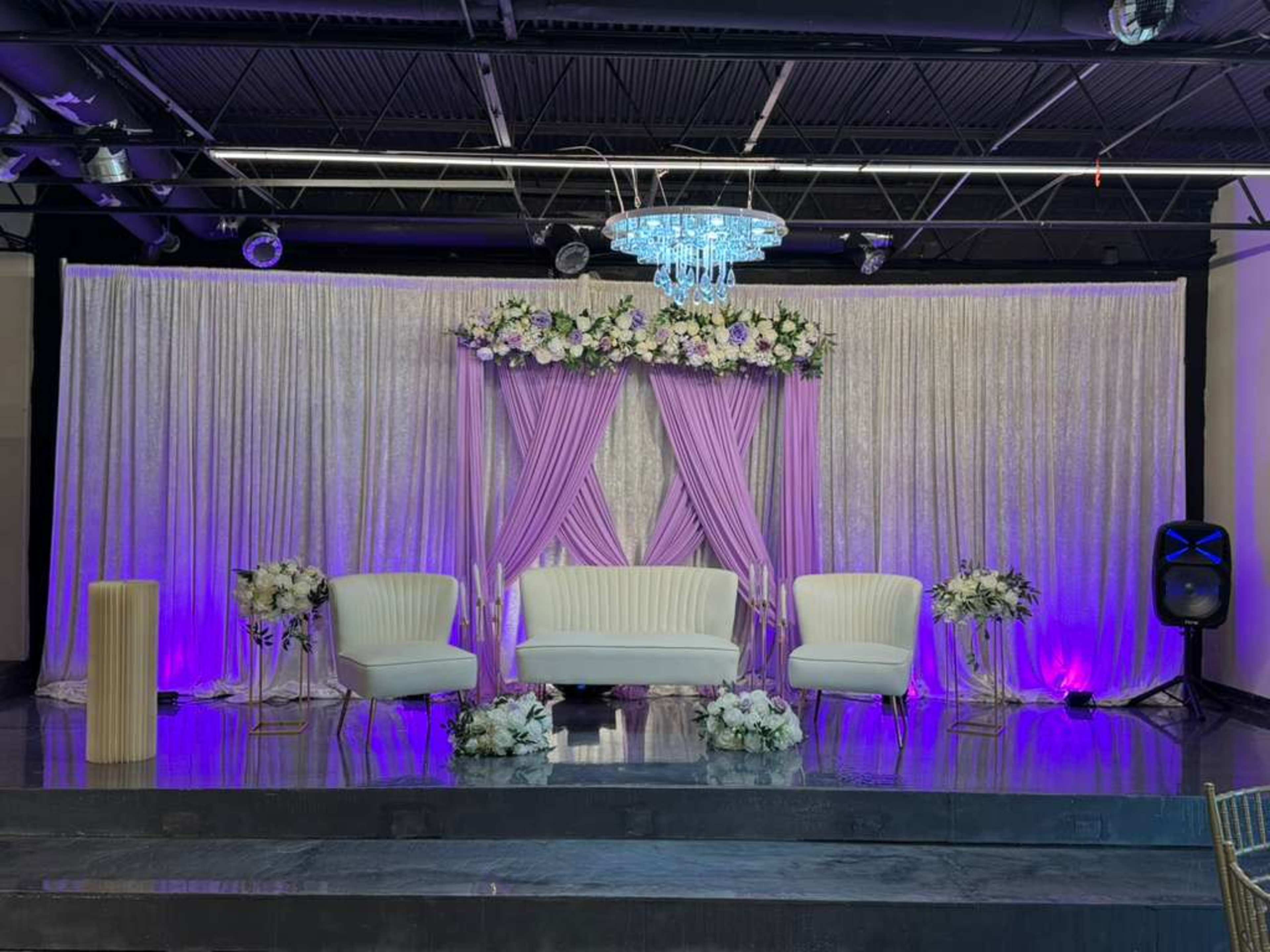 The image depicts a decorated stage for an event, featuring a set of white chairs, floral arrangements, and a chandelier, with purple and silver drapery backdrop.