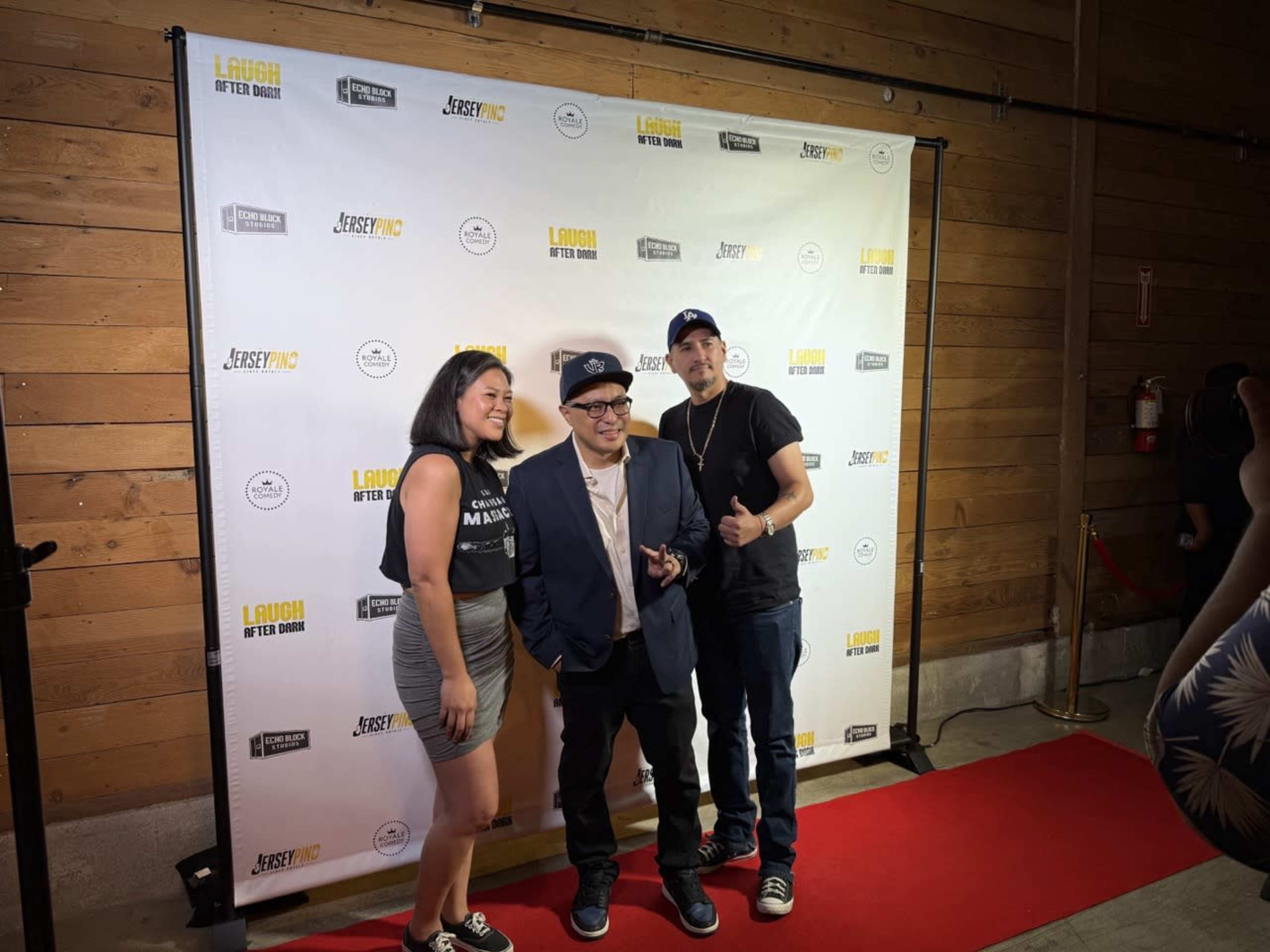 A group of three people, including a man in a suit and two others in casual attire, pose together in front of a branded backdrop at an event.