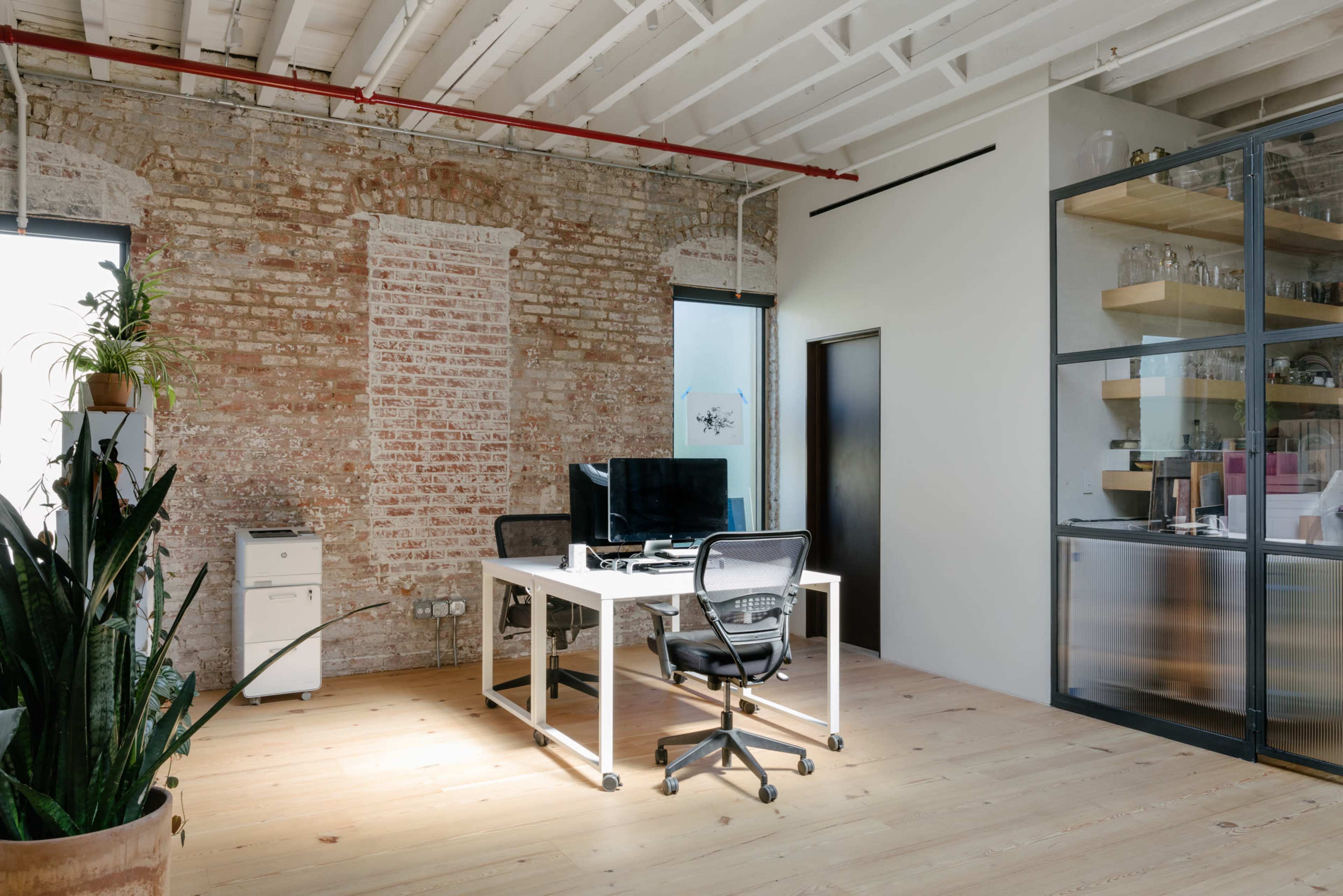 A modern office features a white desk and chair, a computer set up, and brick walls with large windows, alongside a plant and a glass cabinet filled with items.