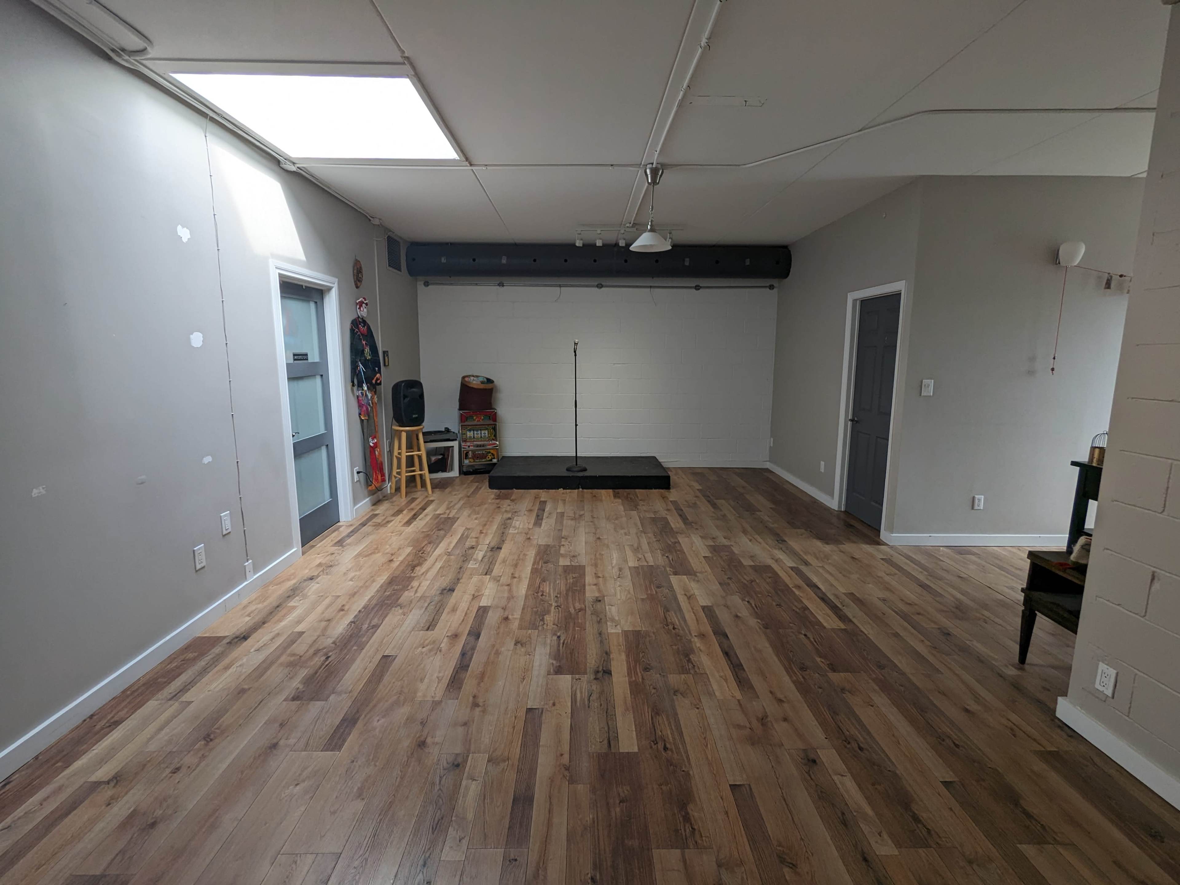 A spacious room with wooden floors, a small stage at one end, and gray walls, featuring a door on the left and another door on the right.