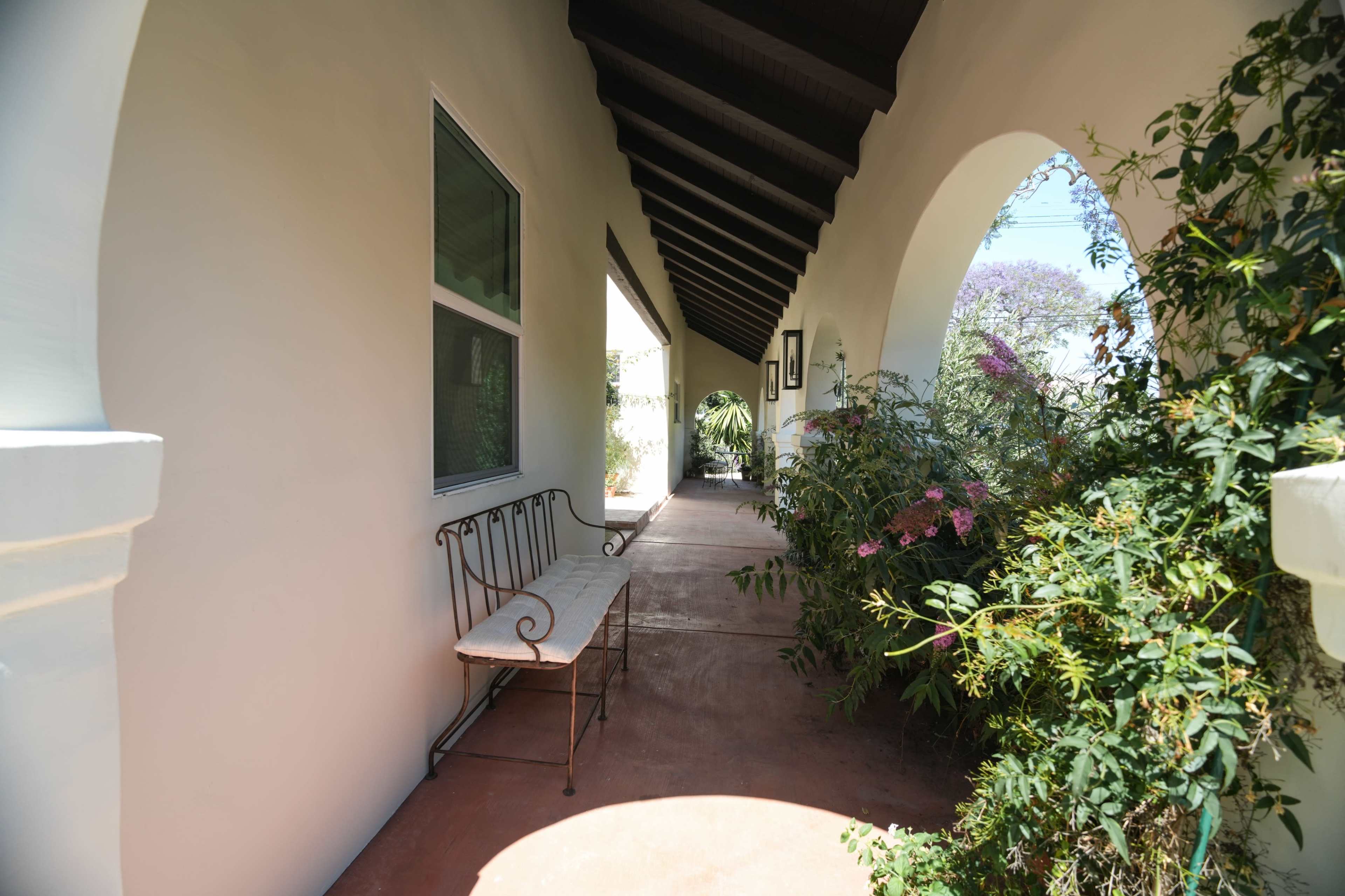 The image shows a covered walkway adorned with a wrought iron bench and surrounded by greenery.