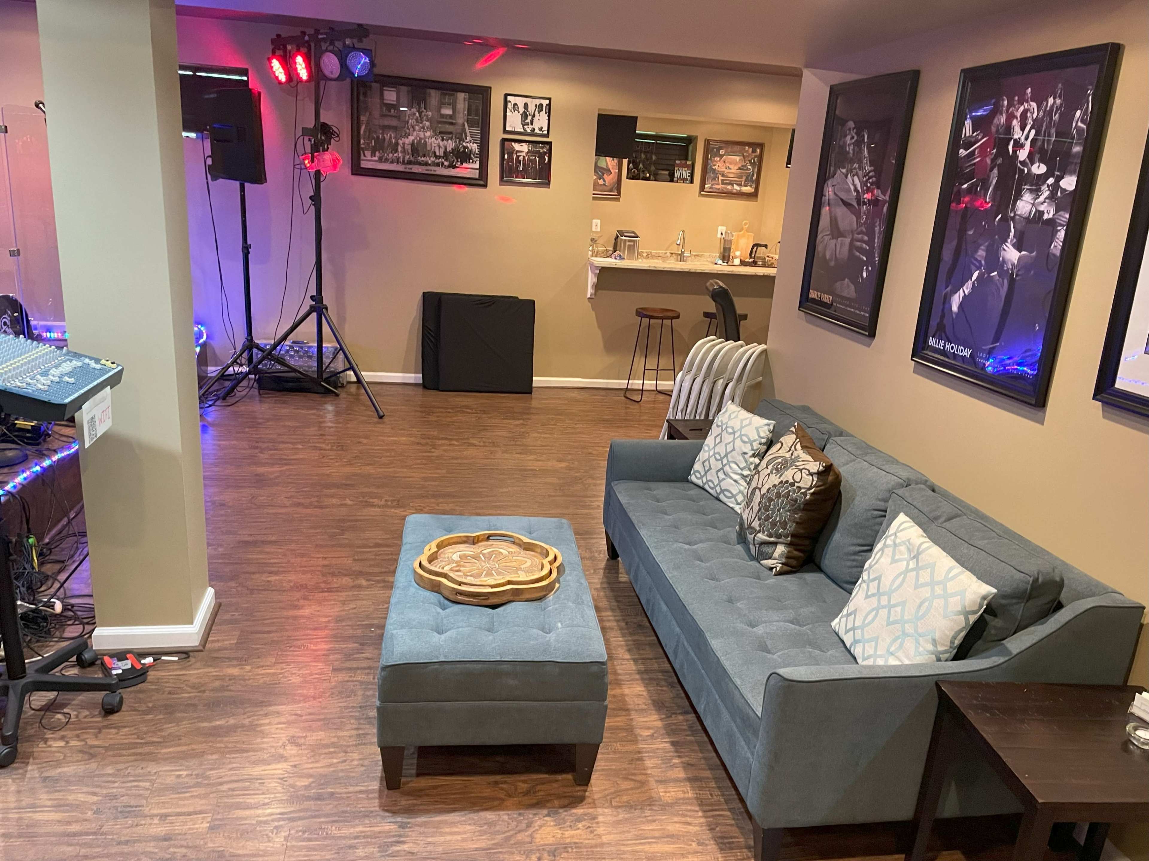 A basement space with a blue couch, an ottoman, a small table, and a music setup with lighting fixtures on one side.