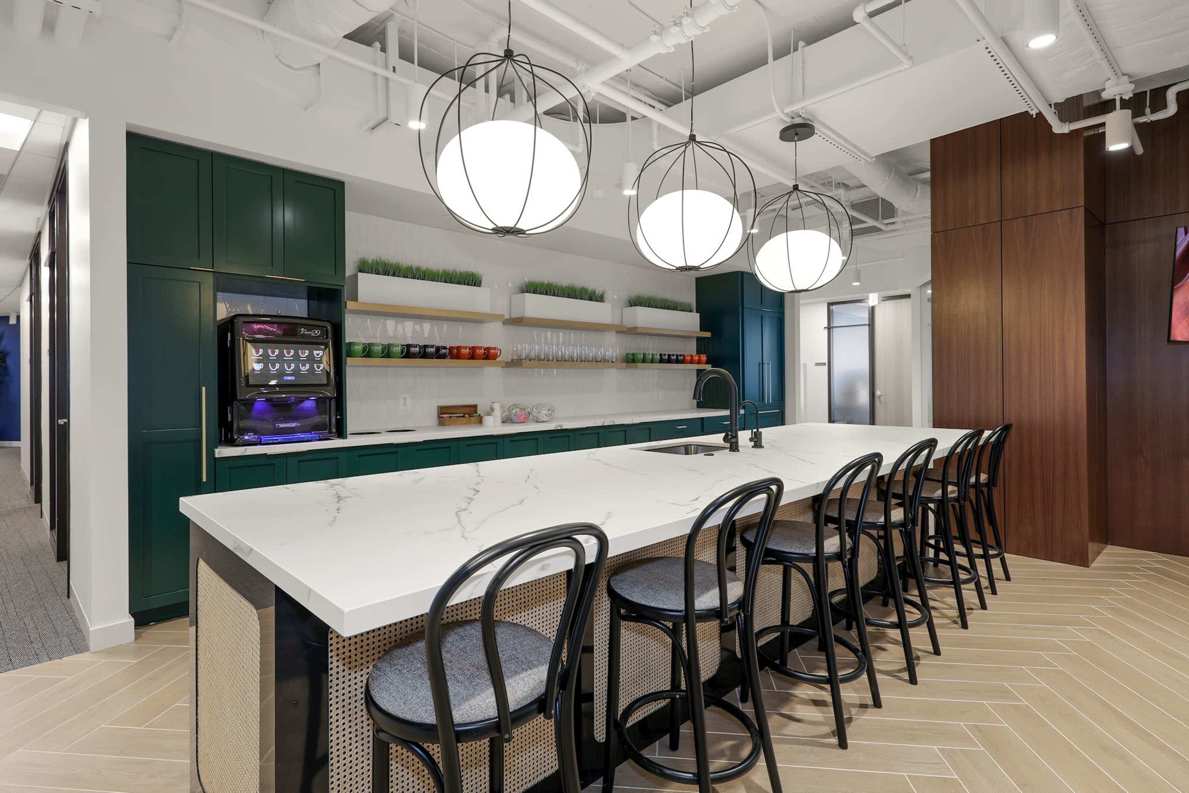 A modern office kitchen with a long marble countertop, black chairs, and pendant lights, complemented by green cabinetry and a drink dispenser.