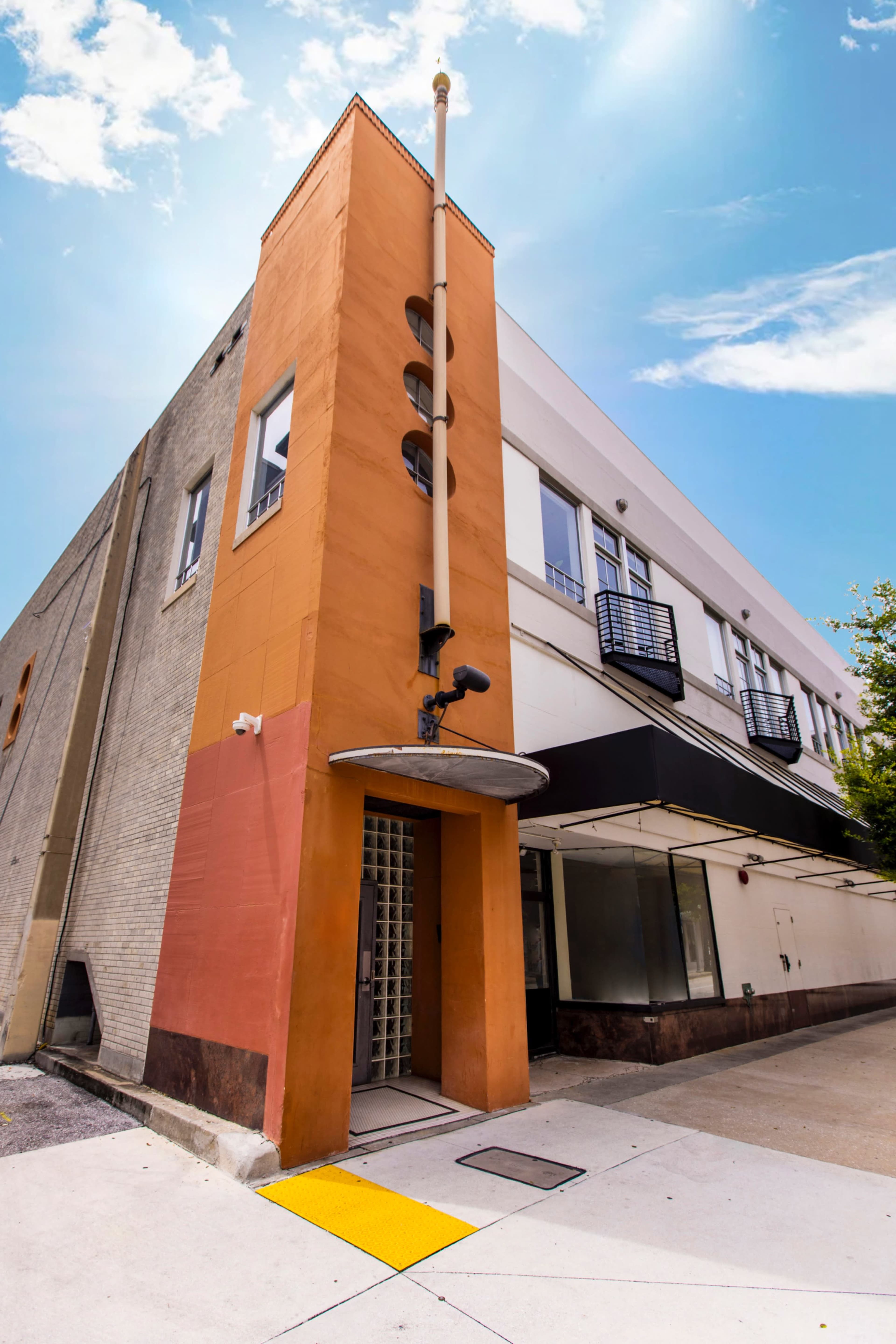The image shows a corner view of a modern, two-story building with an orange and beige exterior, featuring a tall vertical structure and a blue sky in the background.
