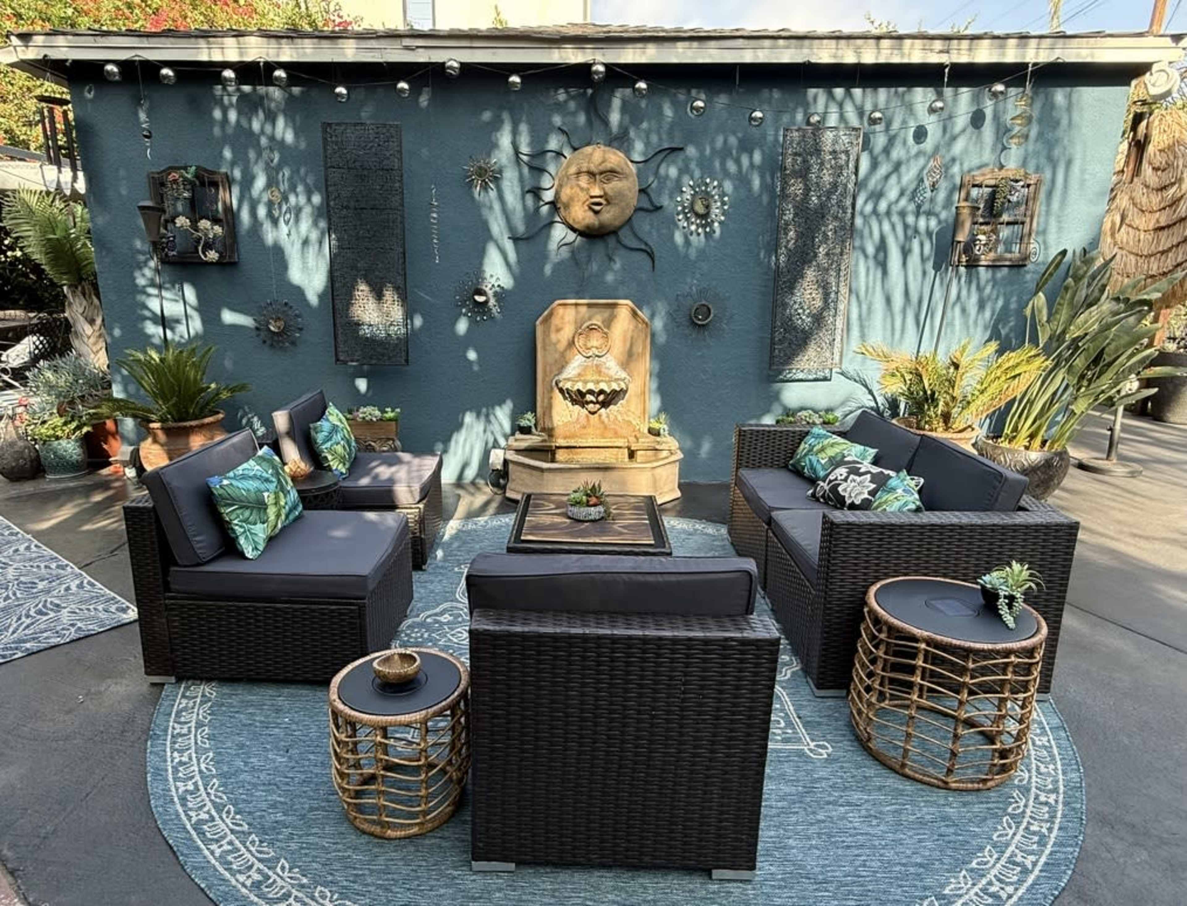A cozy outdoor seating area features dark wicker furniture arranged on a circular patterned rug, with a vibrant blue wall adorned with decorative art and greenery.