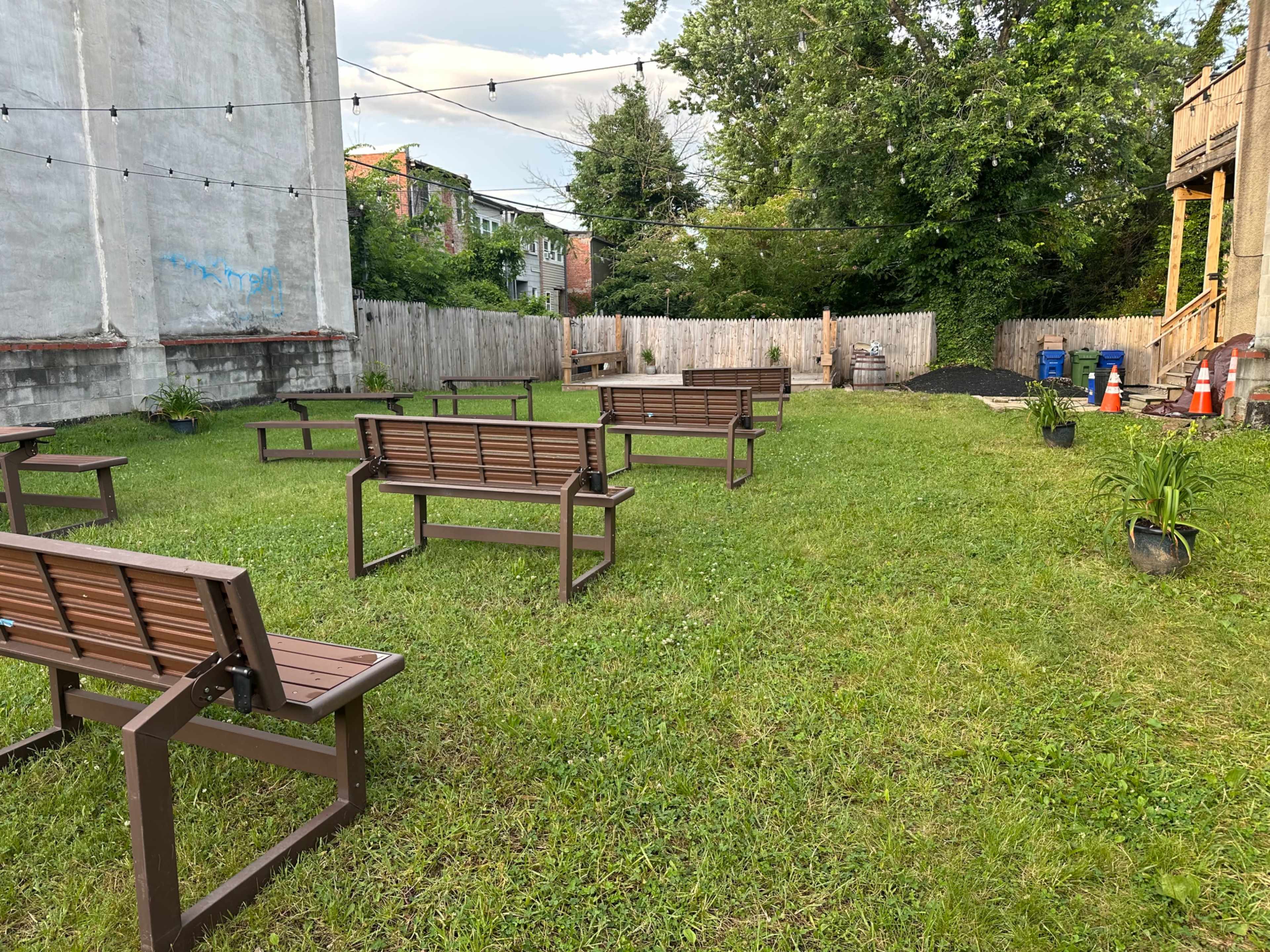 The image shows a grassy area with several metal benches arranged in rows, surrounded by a wooden fence and trees.
