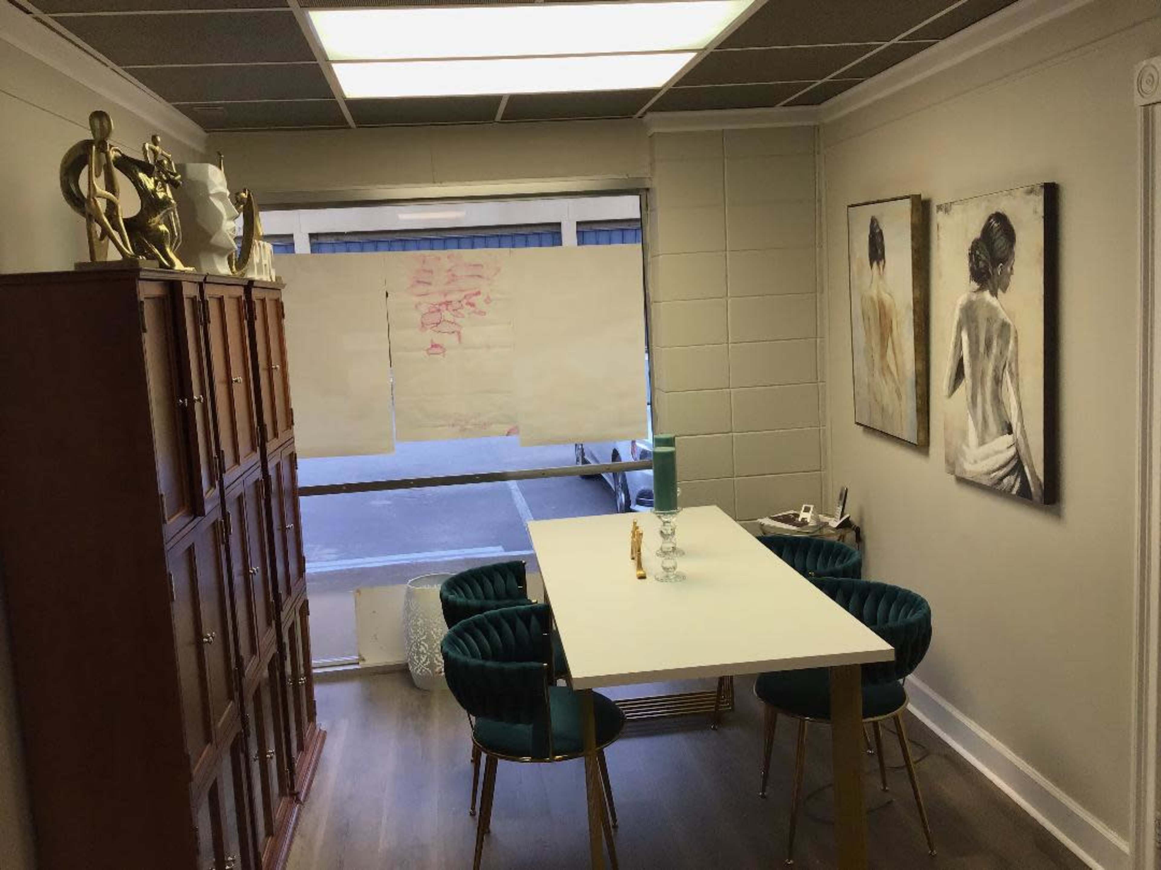 A small office space with a table and four green chairs, a wooden cabinet against the wall, and two framed artworks hanging on the adjacent wall.